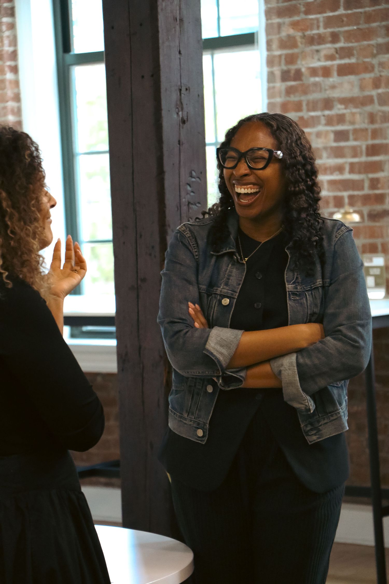Two women laughing together in conversation in brick-walled indoor space.