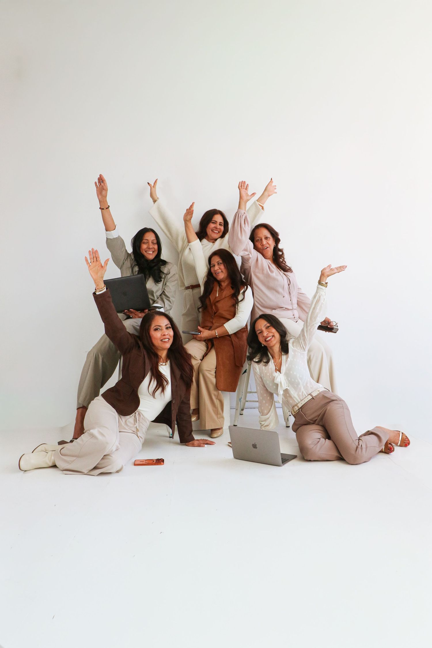 Group of six women celebrating with raised arms and laptops against a white studio background.