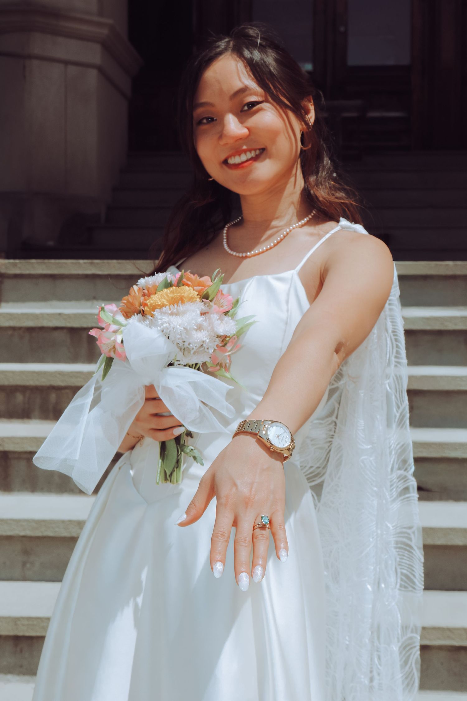 Smiling bride in white dress holding floral bouquet, wearing pearl necklace and gold watch on sunny steps.