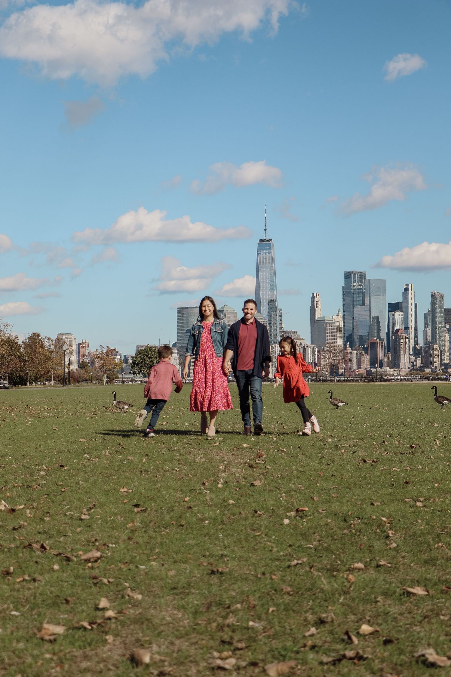 Family of four walks hand-in-hand on grass with NYC skyline and One World Trade Center in background.