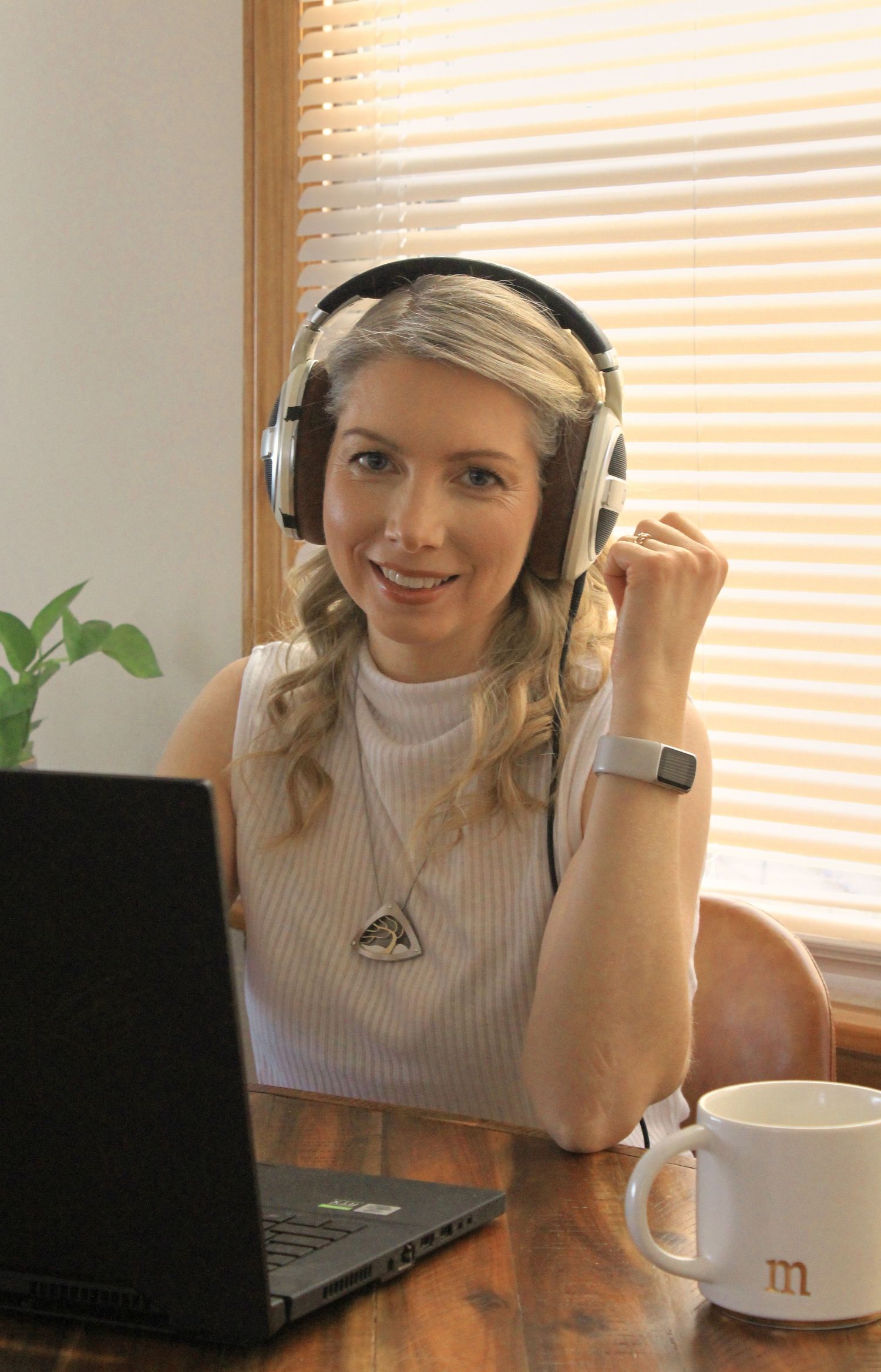 Blonde woman wearing headphones raising fist in victory gesture while working on laptop at desk.