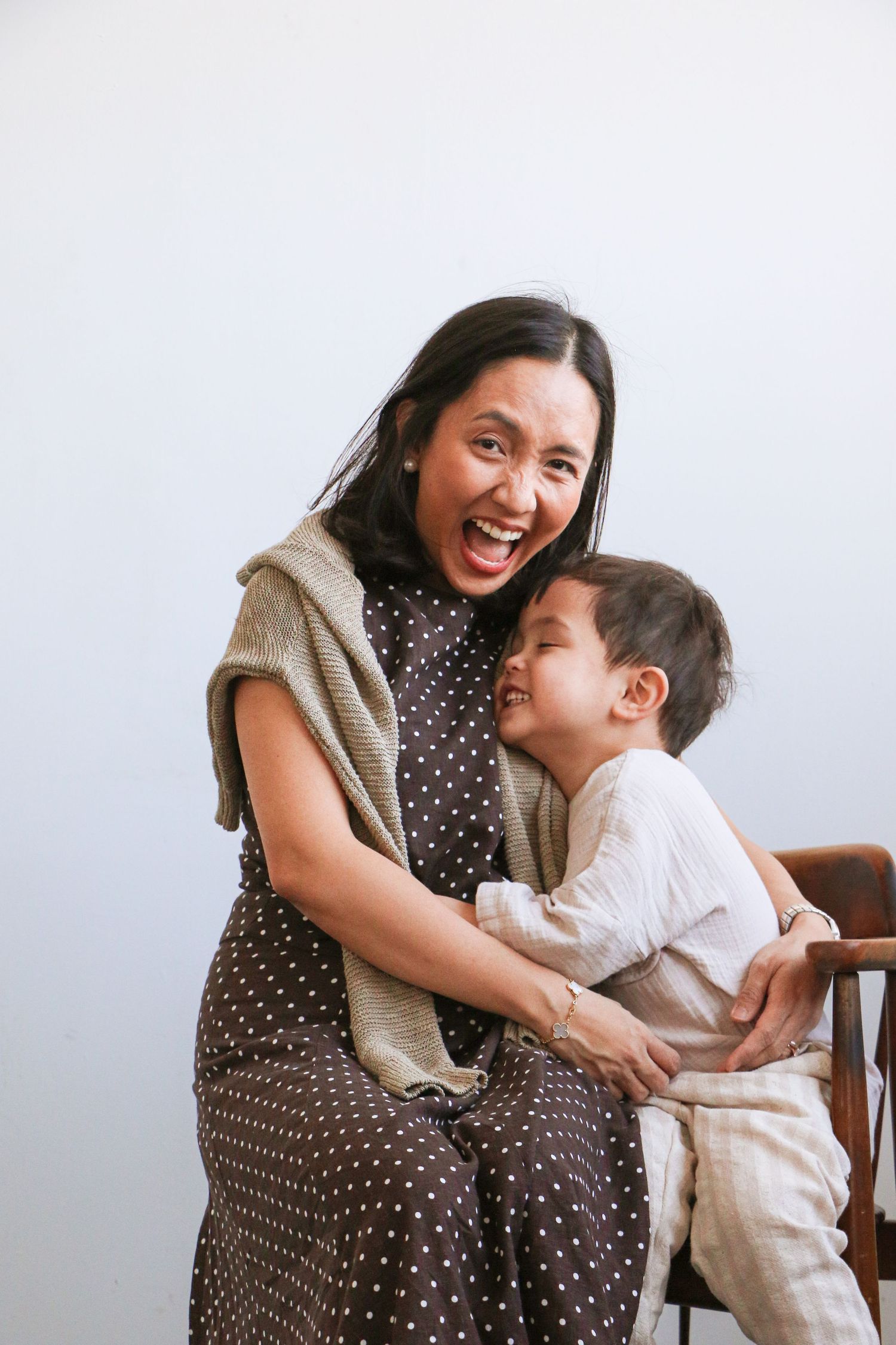 Joyful Asian mother laughing with her toddler son in a brown polka dot dress during a studio photo session.