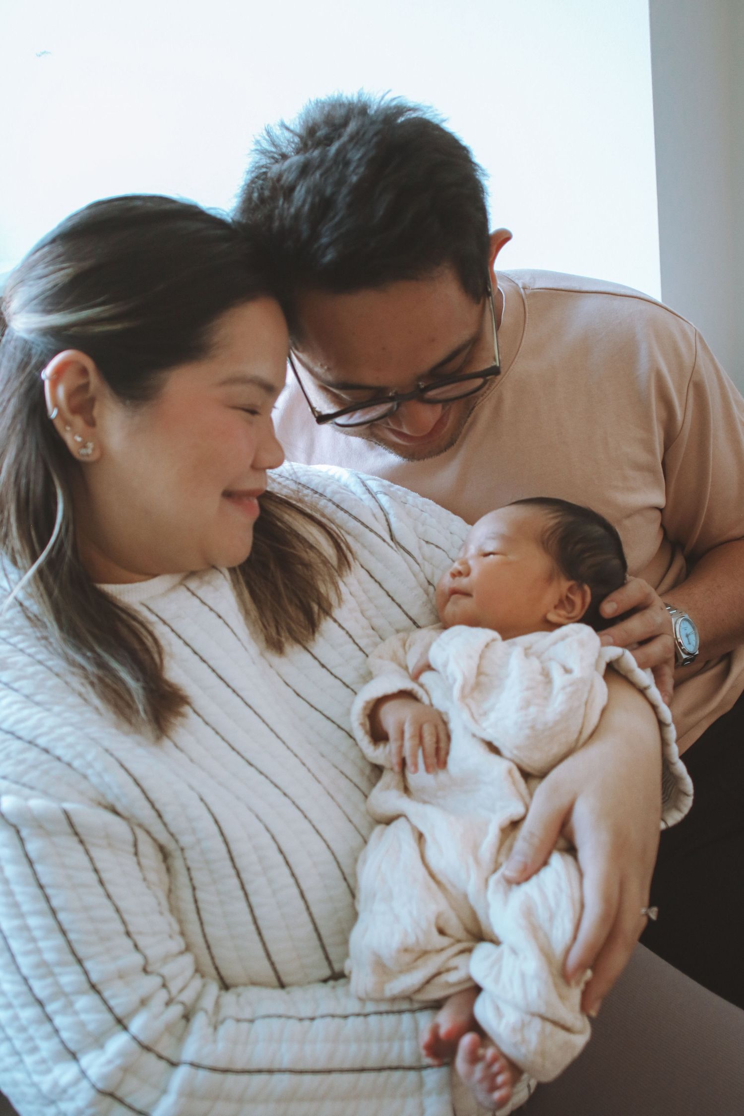 New parents lovingly gaze at sleeping newborn wrapped in white swaddle blanket indoors.
