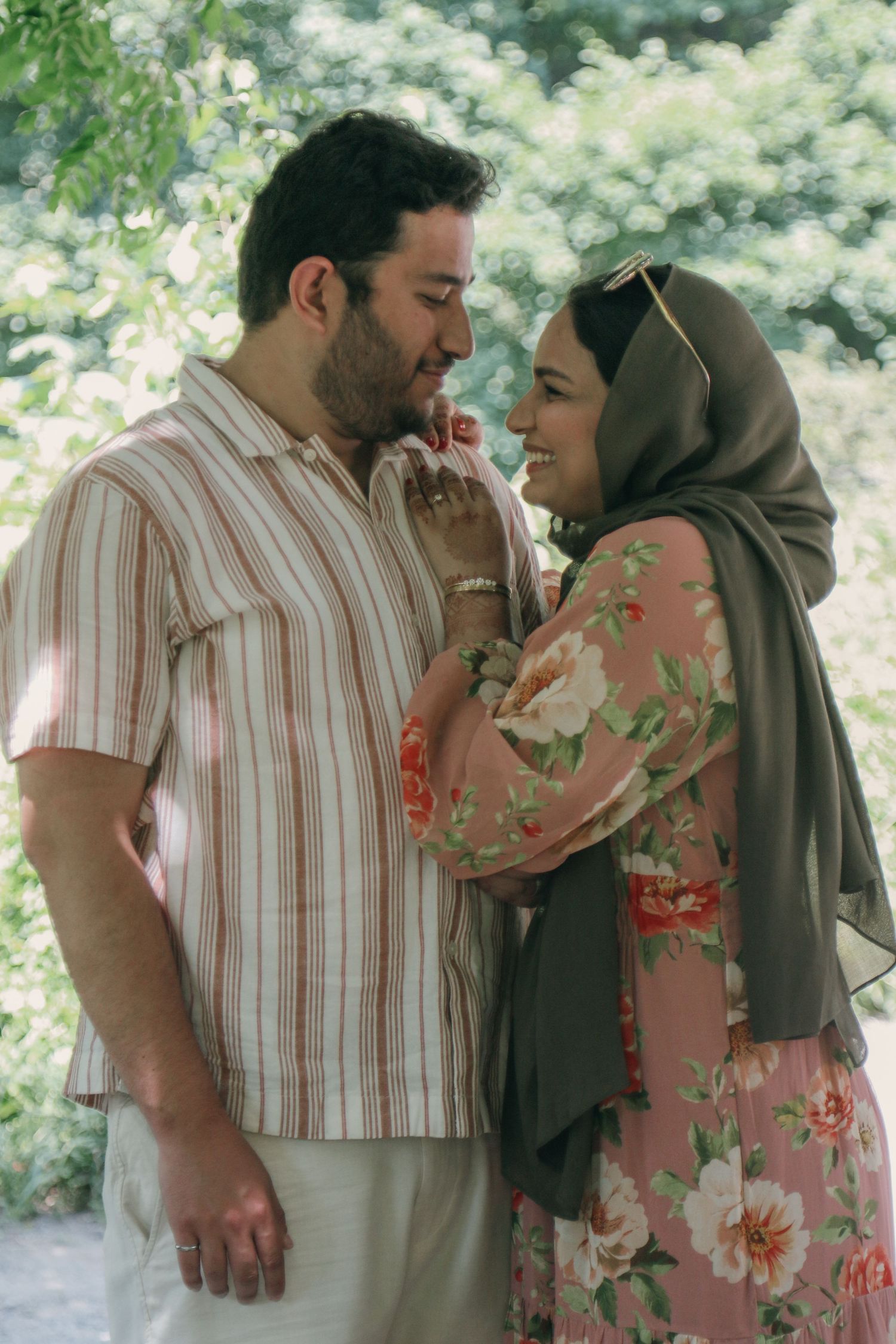 Smiling couple embracing outdoors among green trees, the man in a striped shirt and the woman in an olive hijab and floral dress.