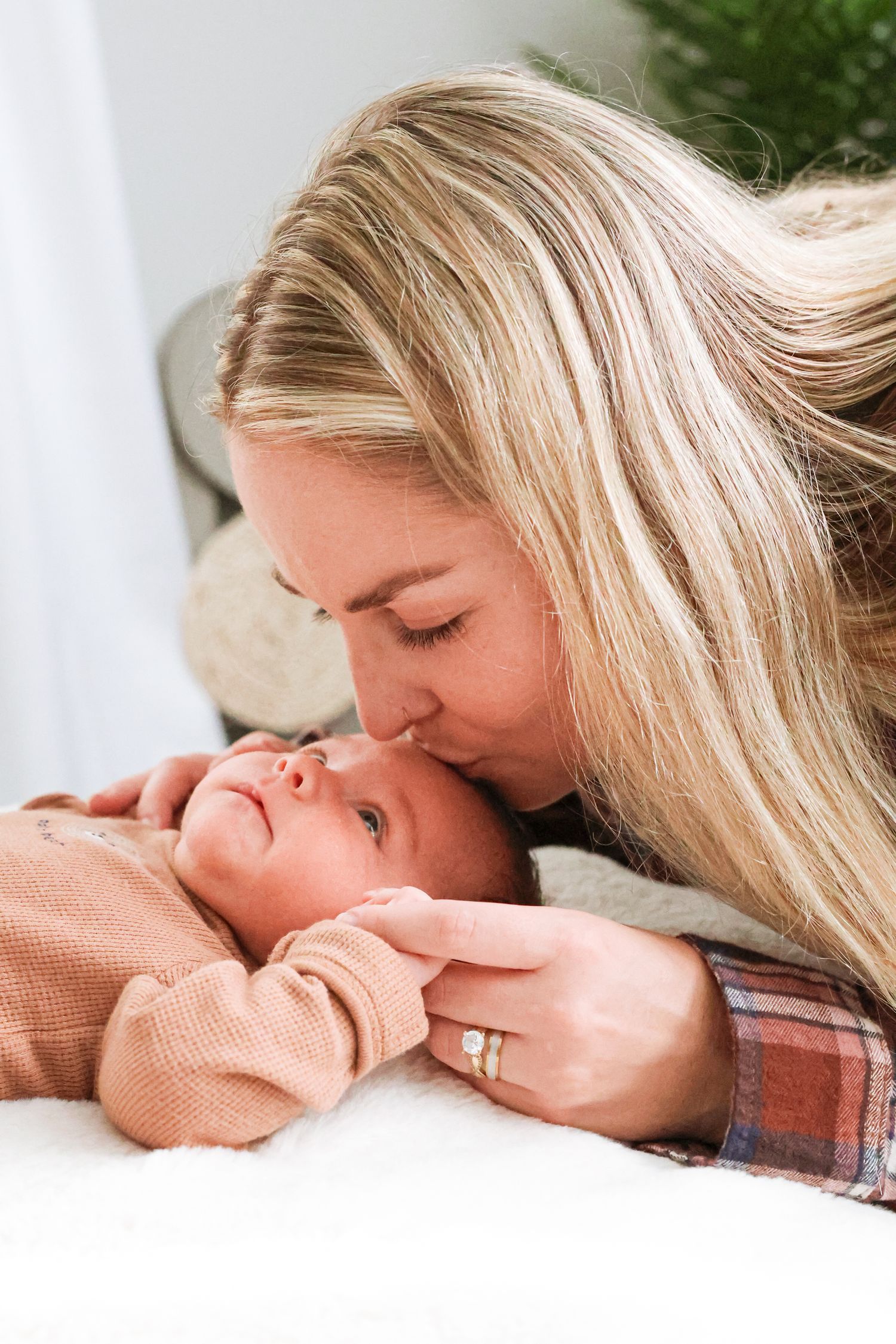 A blonde mother in a plaid shirt gently kisses the forehead of her newborn baby, who rests on a soft white blanket.