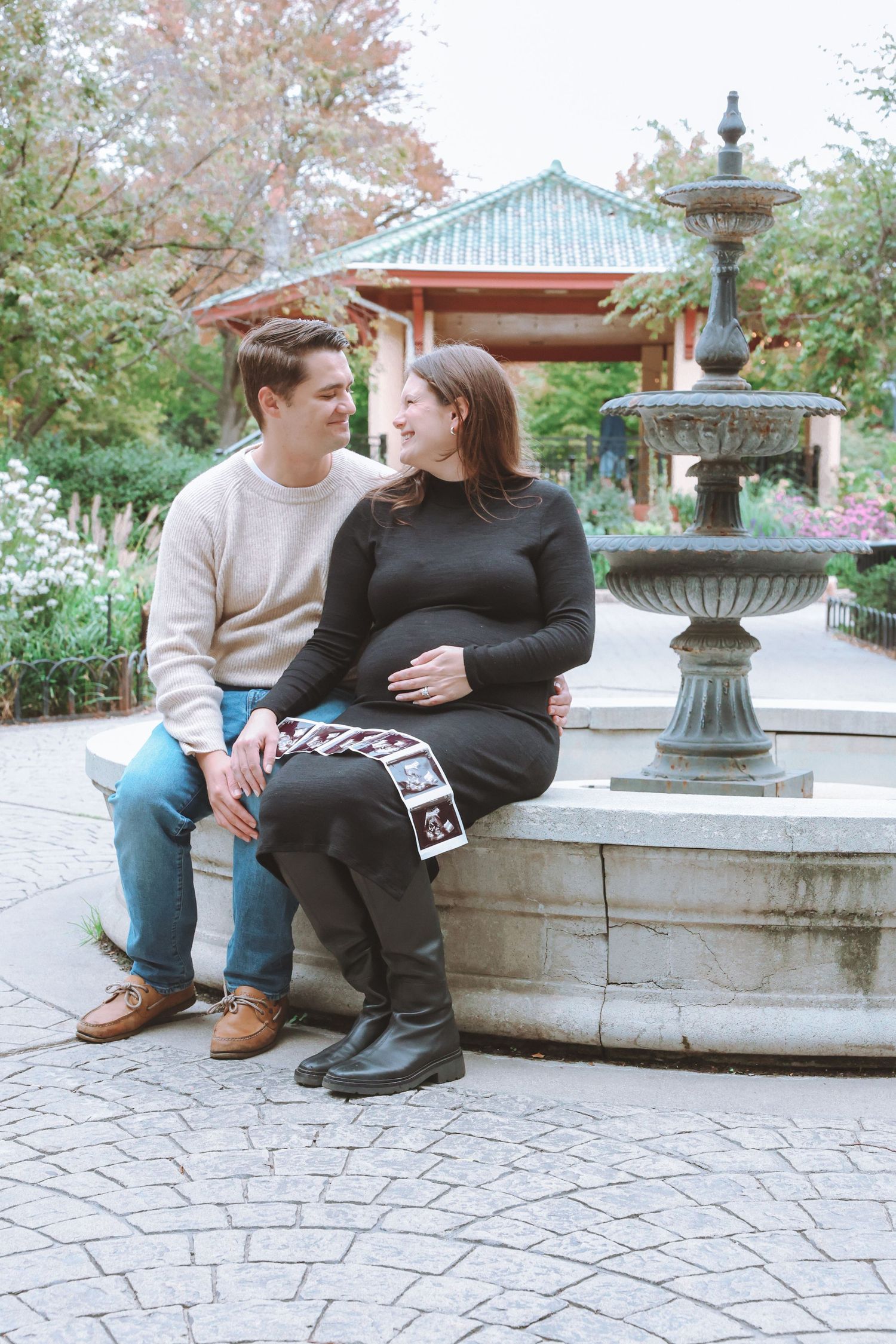 Expecting couple sits on a stone fountain in a garden, the pregnant woman holding an ultrasound photo near a pagoda-style pavilion.