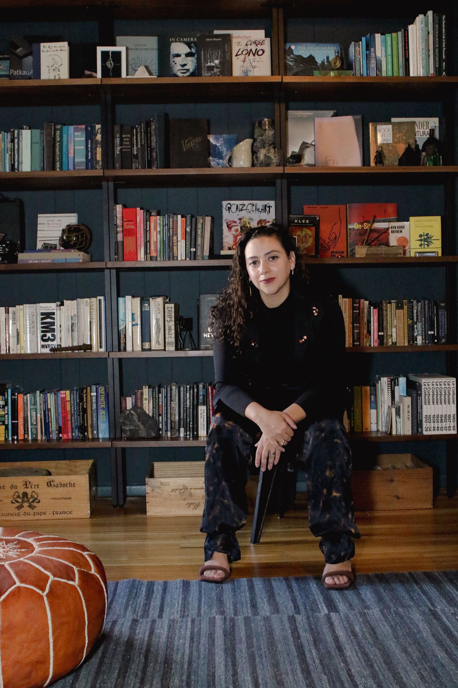 Woman with curly hair sitting on stool in front of large dark bookshelf filled with books.