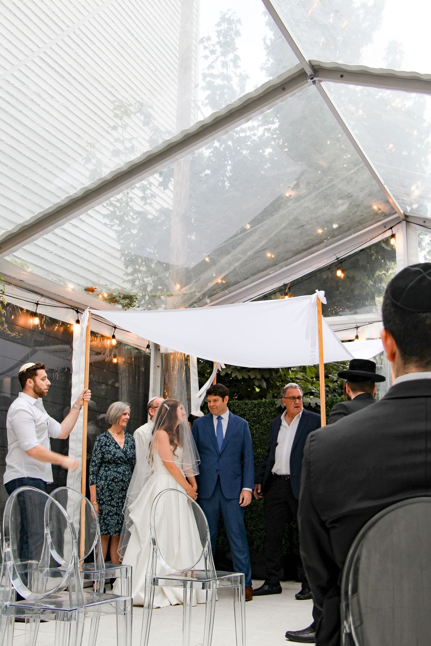 Jewish wedding ceremony under a chuppah inside a clear tent with string lights and guests seated.