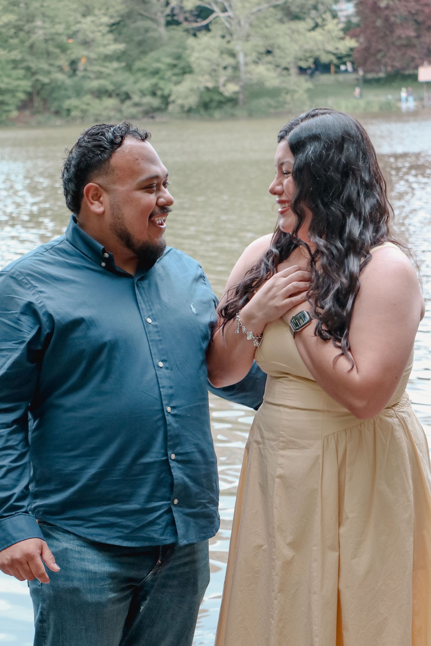 Couple smiling at each other near a lake, man in teal shirt, woman in gold dress, trees in background.