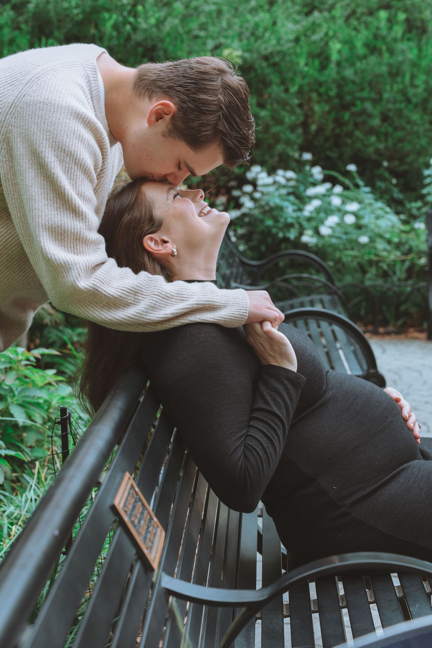 A man in a cream sweater kisses the forehead of a smiling pregnant woman in black as she leans back on a park bench among greenery.