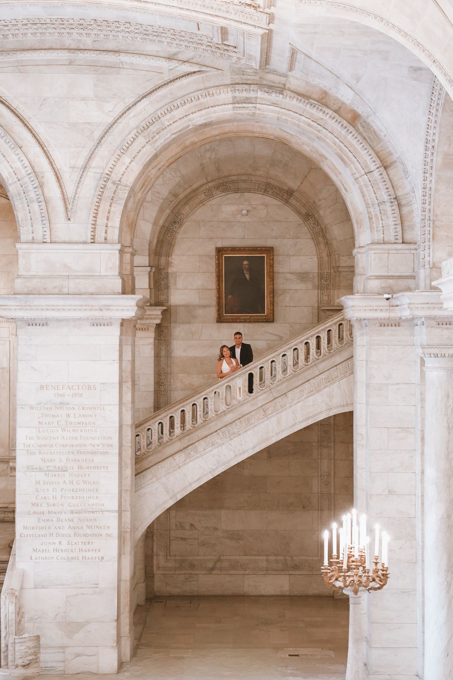 Couple in wedding attire pose on a grand marble staircase beneath soaring arches, with a framed portrait and a glowing chandelier nearby.