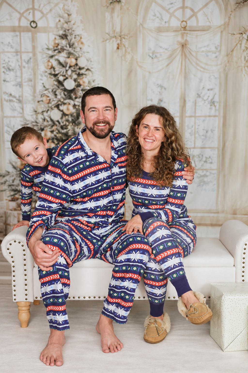 Family of three wearing matching blue holiday pajamas sitting on a white couch in front of a decorated Christmas tree.