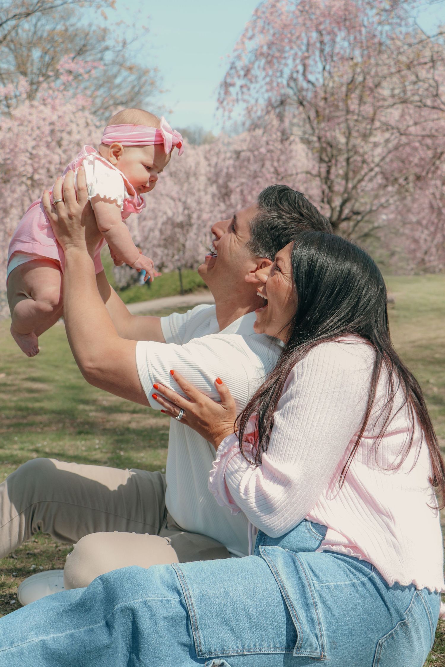 Happy couple sitting outdoors lifting smiling baby girl under pink cherry blossom trees in spring.