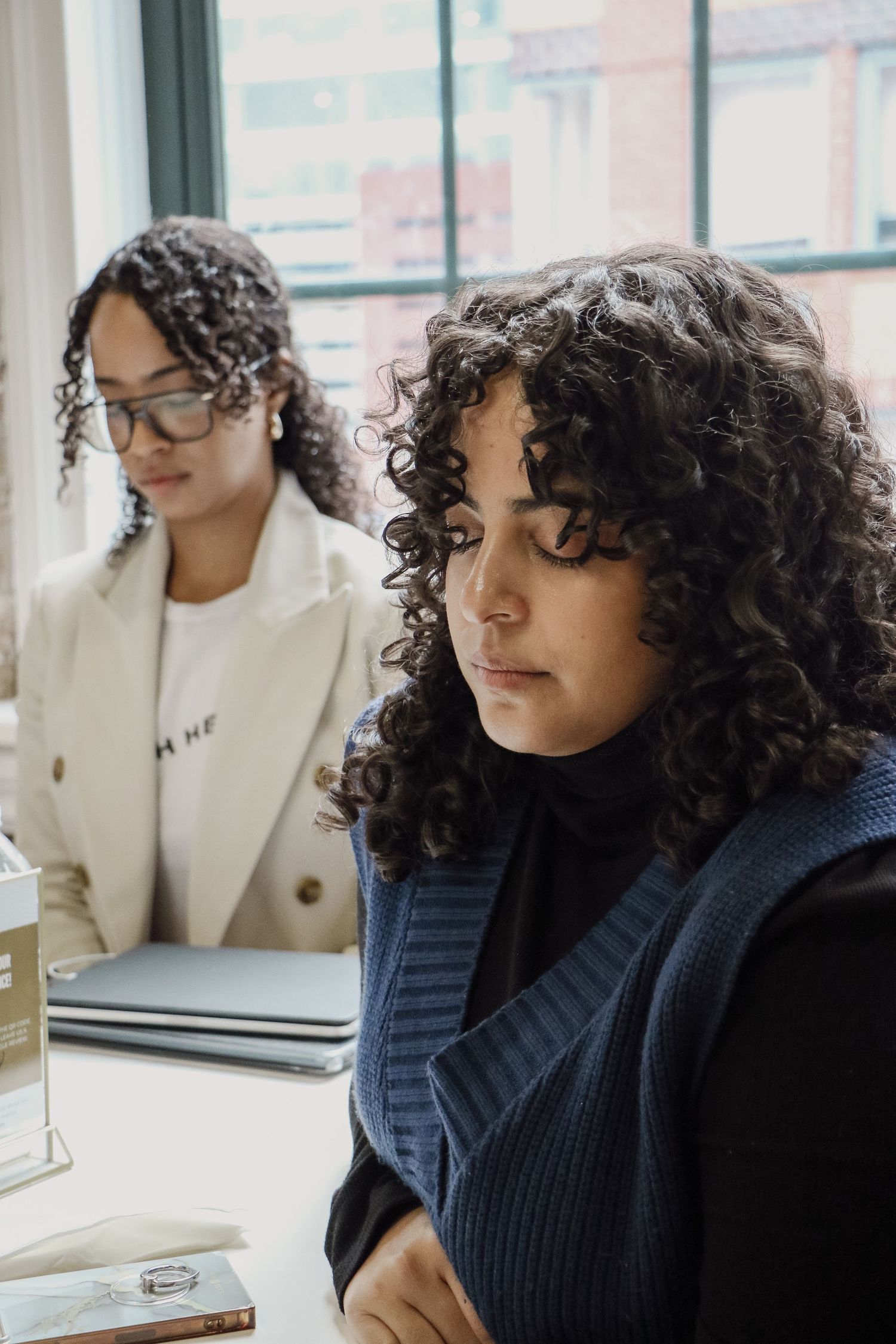Two curly-haired women in professional attire focused at a bright office workspace.