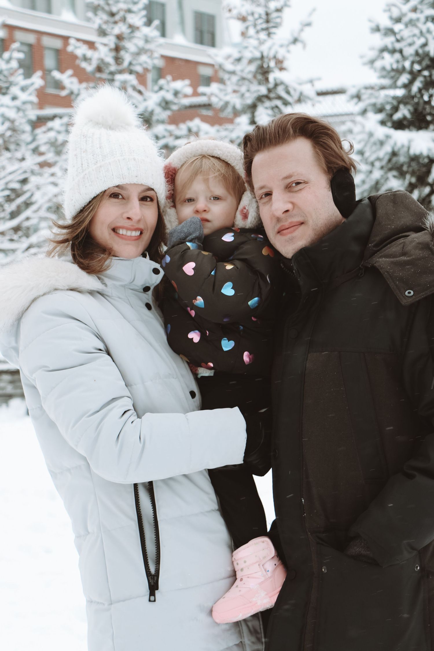 Family with a baby posing outdoors in a snowy winter setting near snow-covered trees.