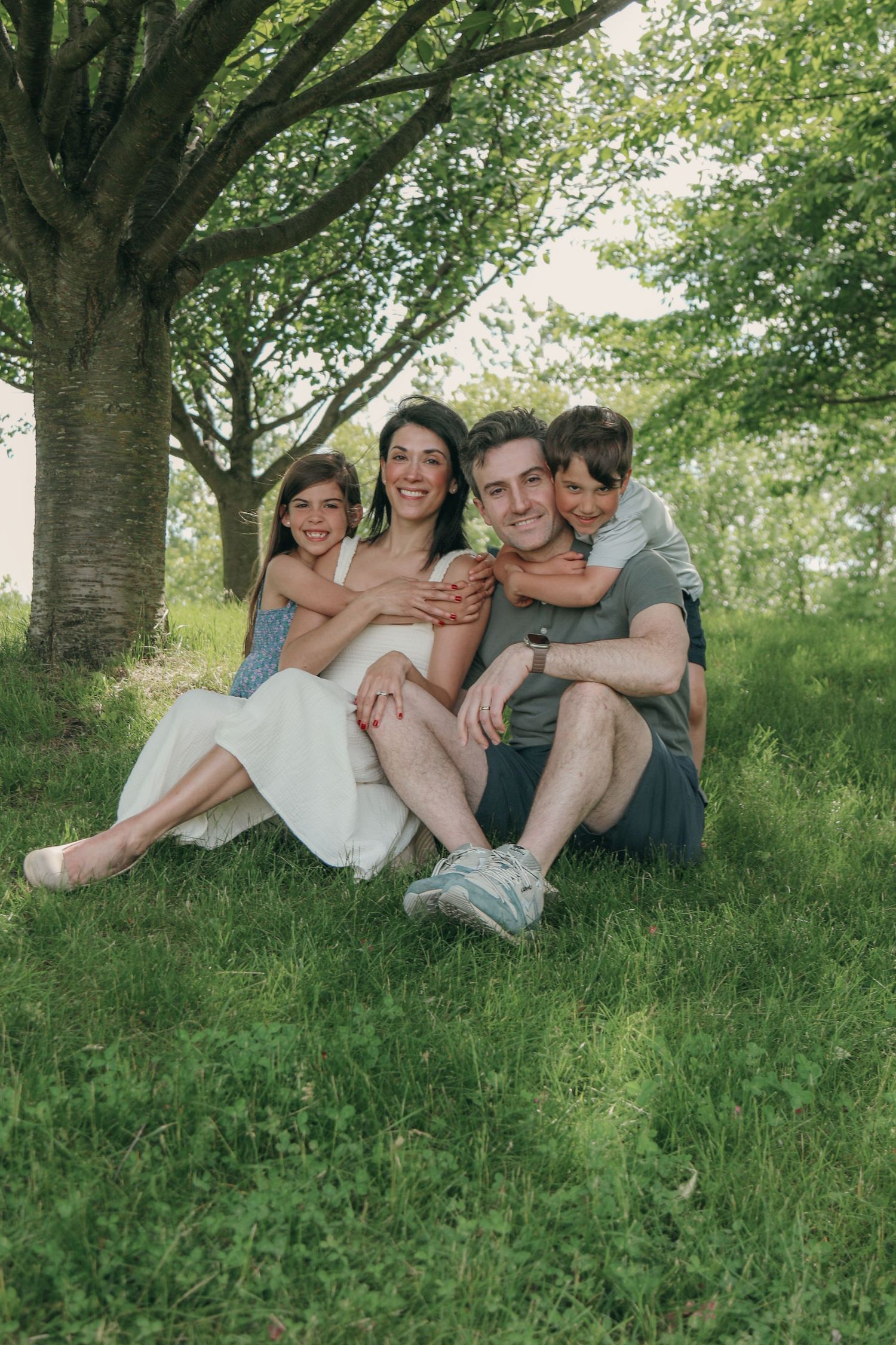 Happy family of four sitting on green grass under a large tree, smiling and embracing in a lush outdoor setting.