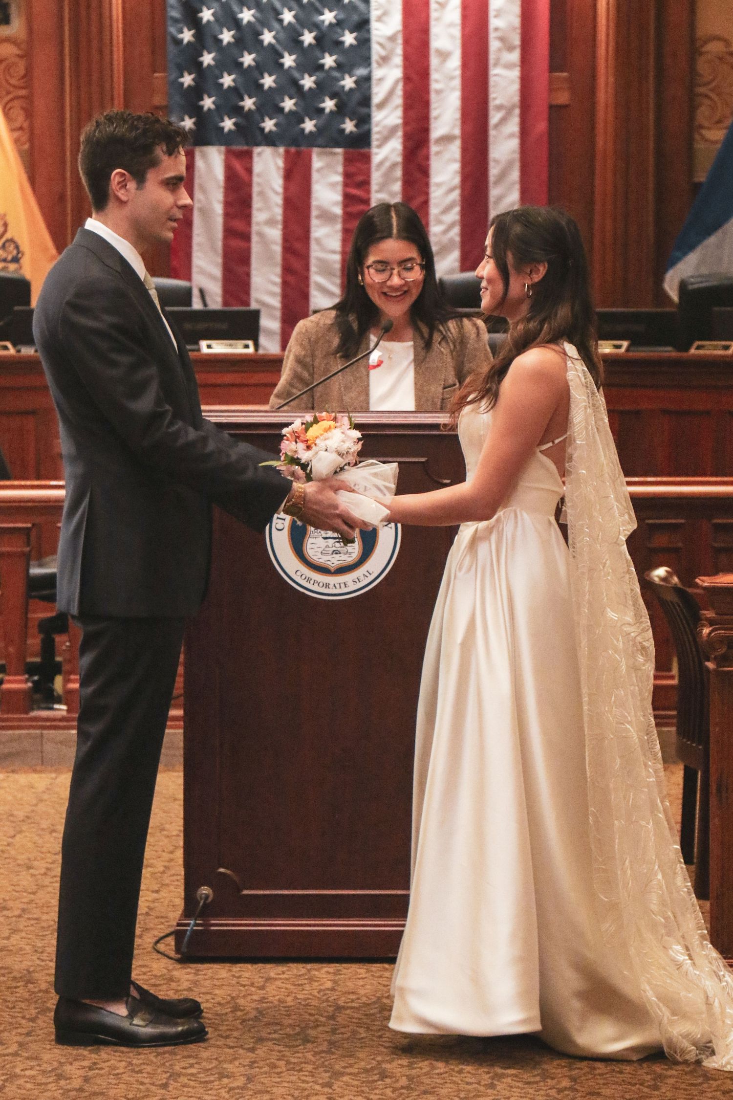 Couple exchanging vows at a civil wedding ceremony before an officiant, with an American flag backdrop.