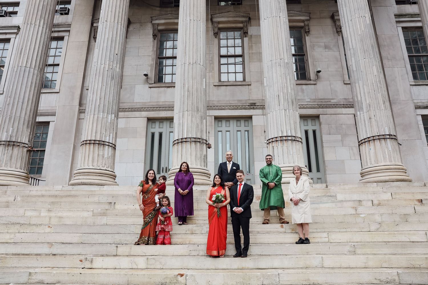 Wedding party in colorful formal attire poses on the wide stone steps of a neoclassical building with tall columns.