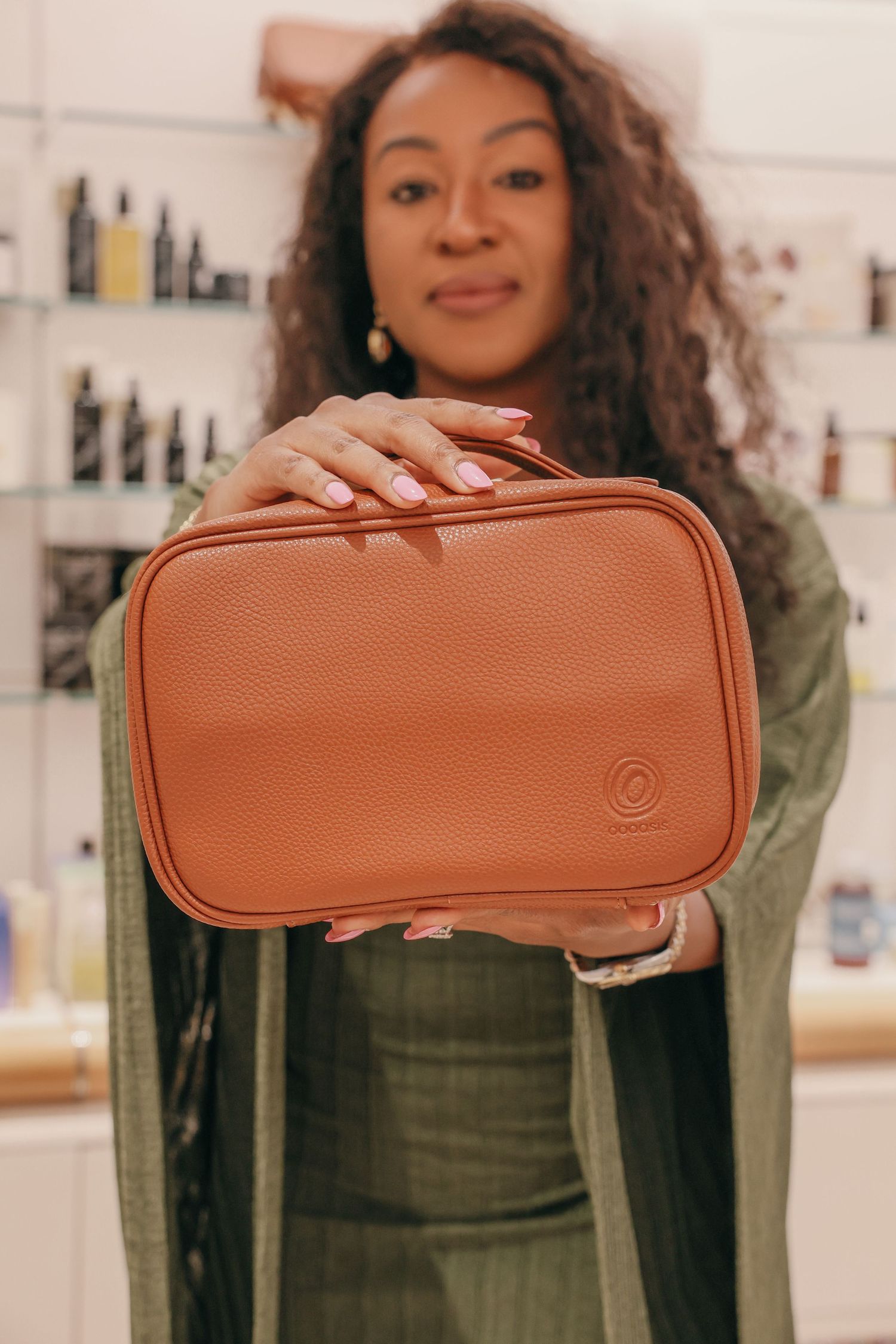 Smiling woman with curly hair holds a tan leather toiletry bag with a embossed logo in front of shelves of skincare bottles.