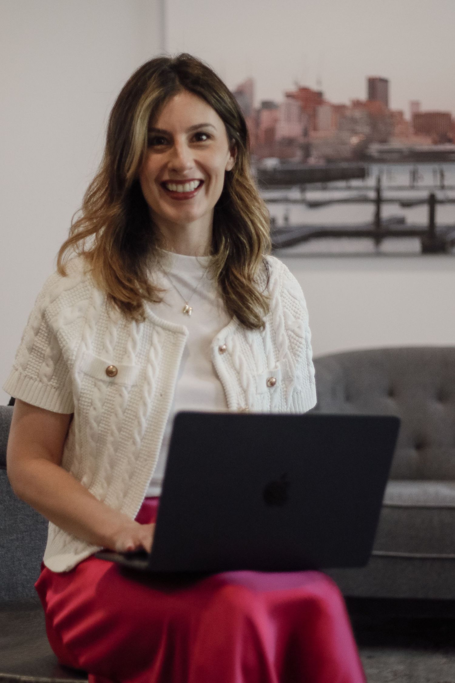 Smiling woman in cream knit vest holding MacBook laptop while seated in modern office space.