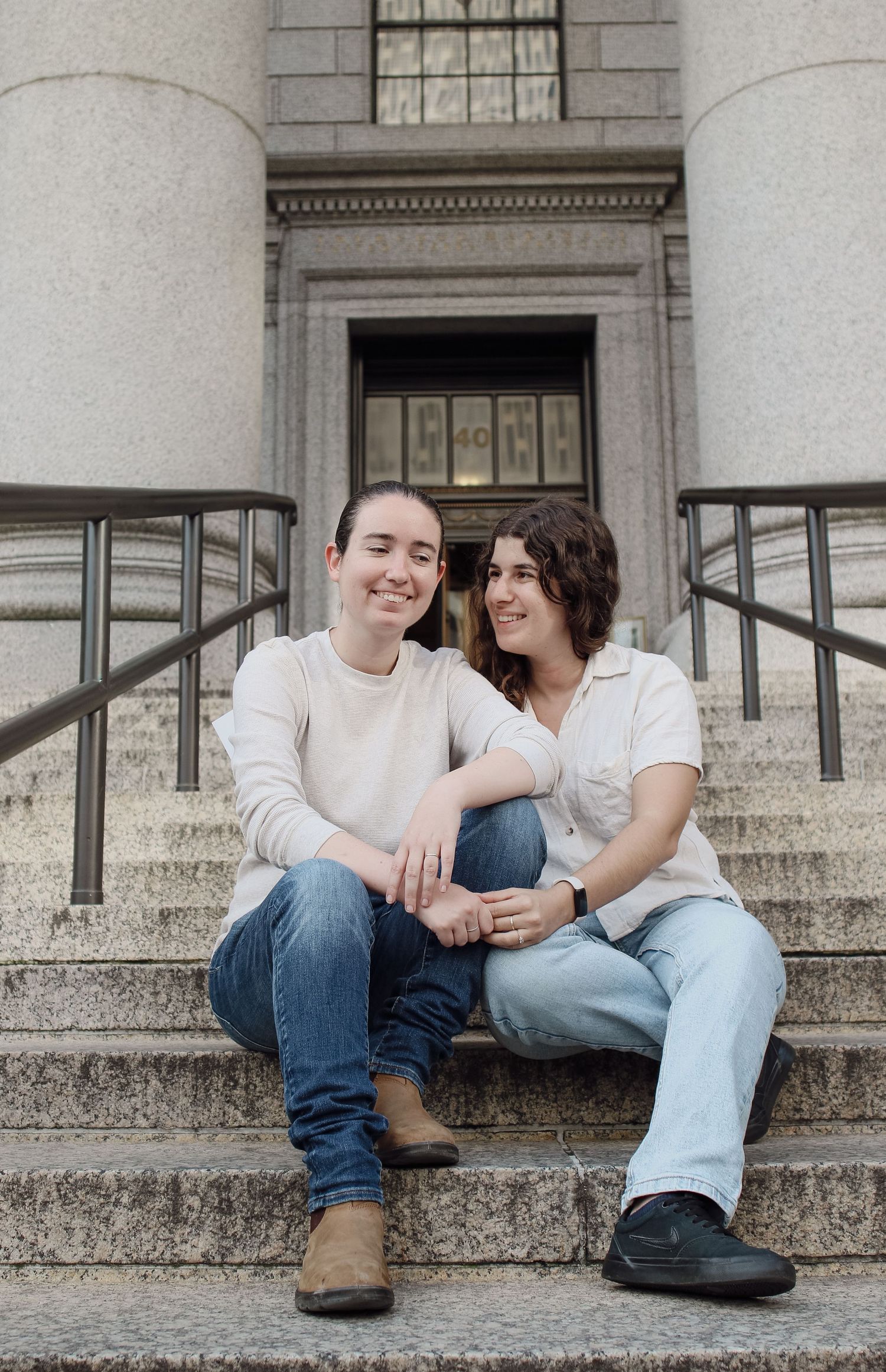 Two women sitting together on stone steps of a classic building, smiling and holding hands.