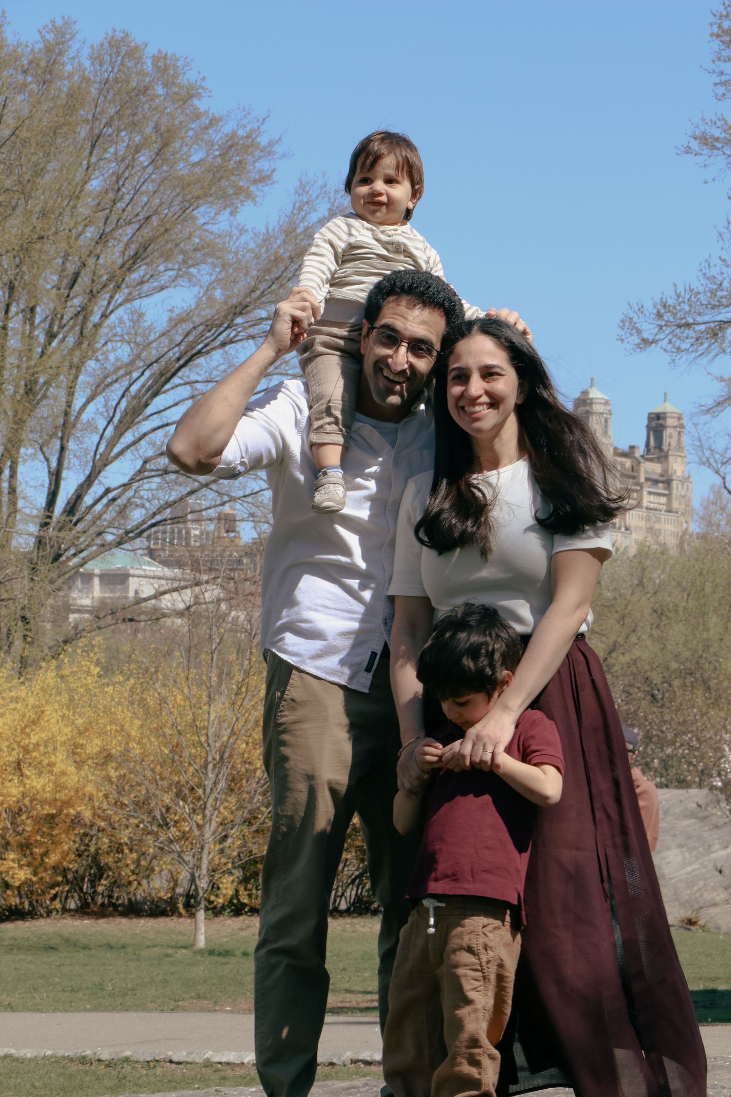 Family of four posing in Central Park, dad holding toddler on shoulders, mom hugging older child, spring trees behind.