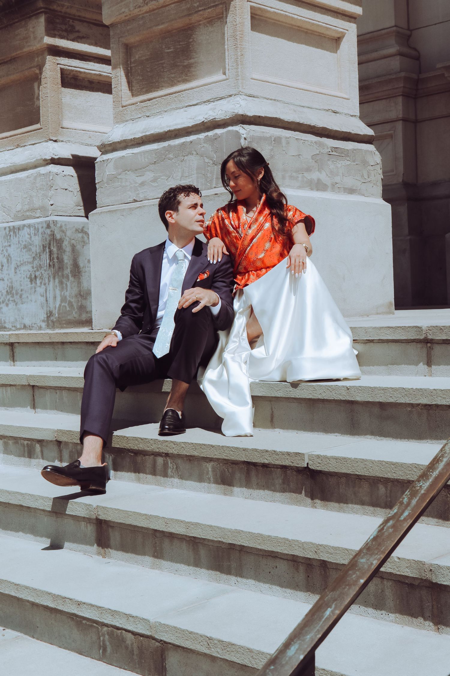 A couple in formal attire sits on stone steps, the woman in a red and white gown gazing at the suited man.
