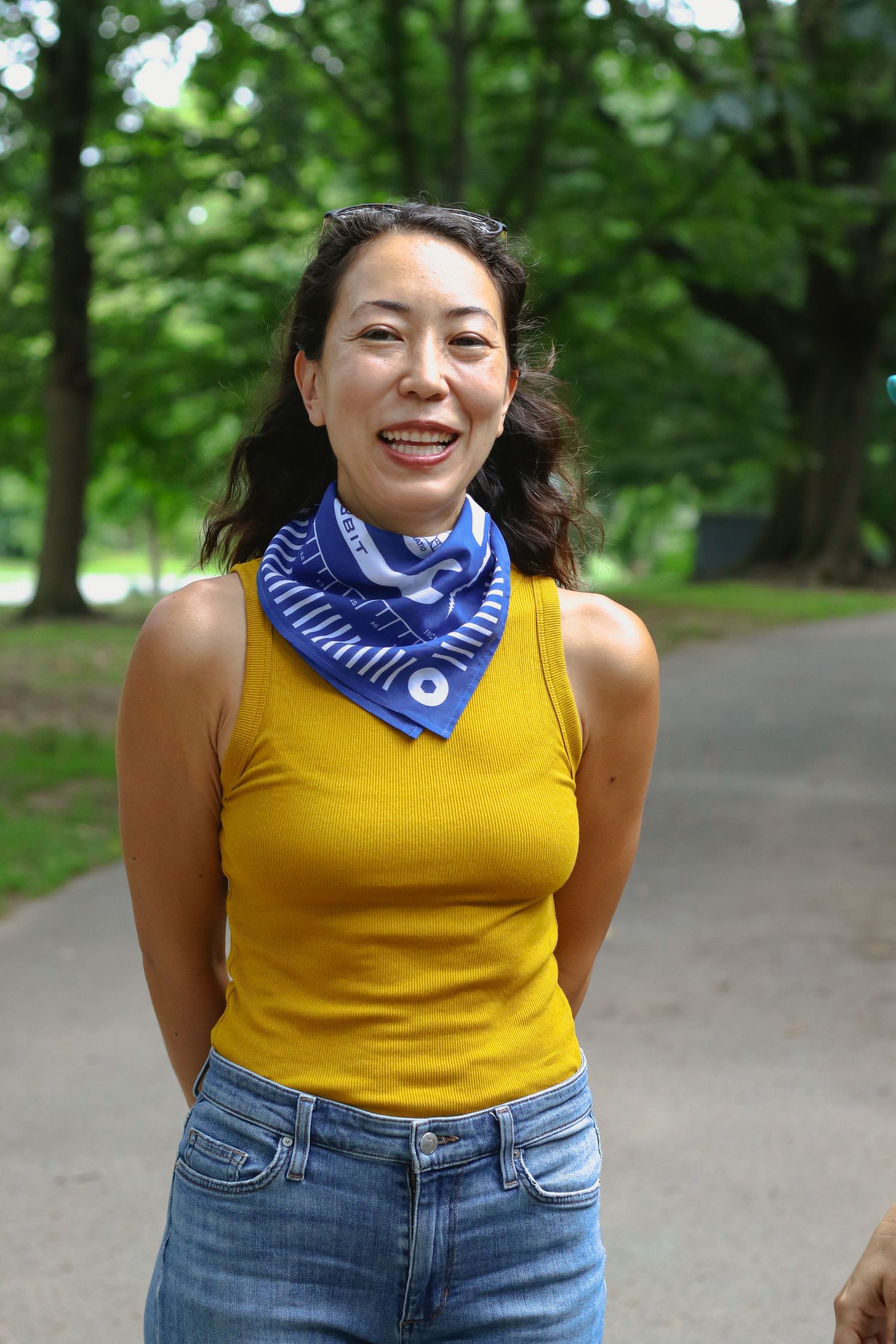 Woman in yellow tank top with blue bandana scarf smiling in tree-lined park path.
