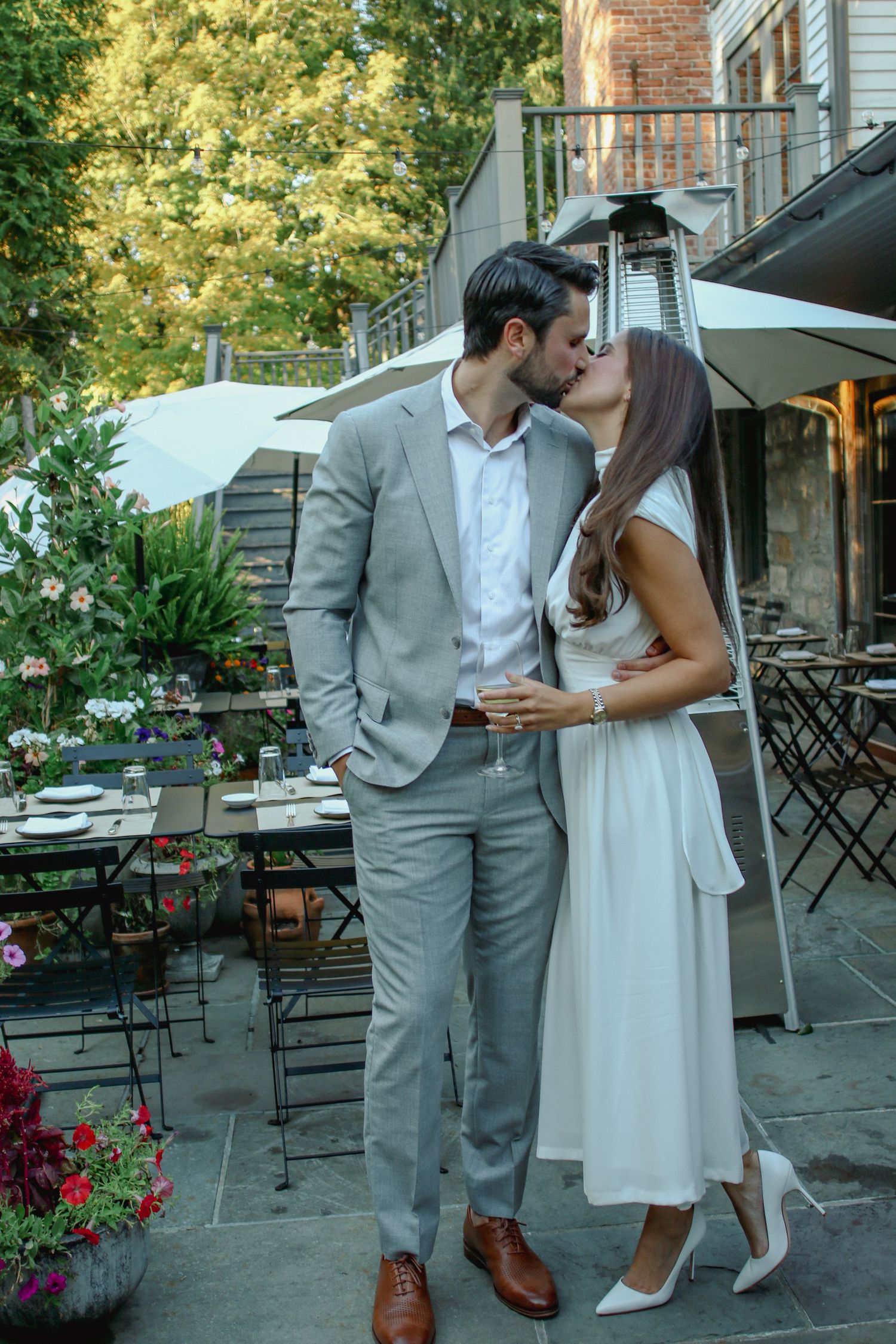 Couple sharing a romantic kiss on a flower-filled outdoor patio, dressed in grey suit and white dress.