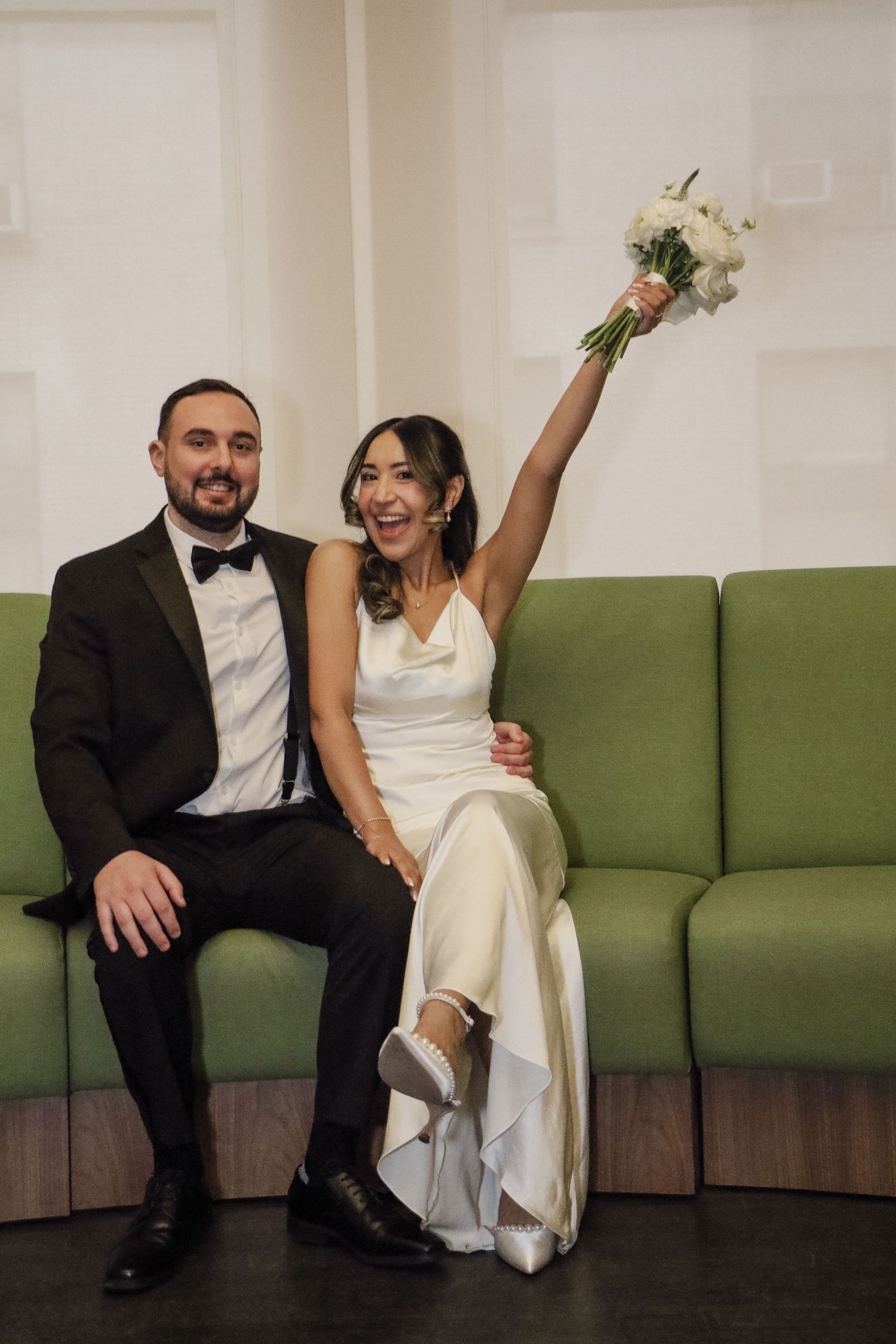 Jubilant bride in white satin gown raises bouquet while seated with groom in black tuxedo on green sofa.