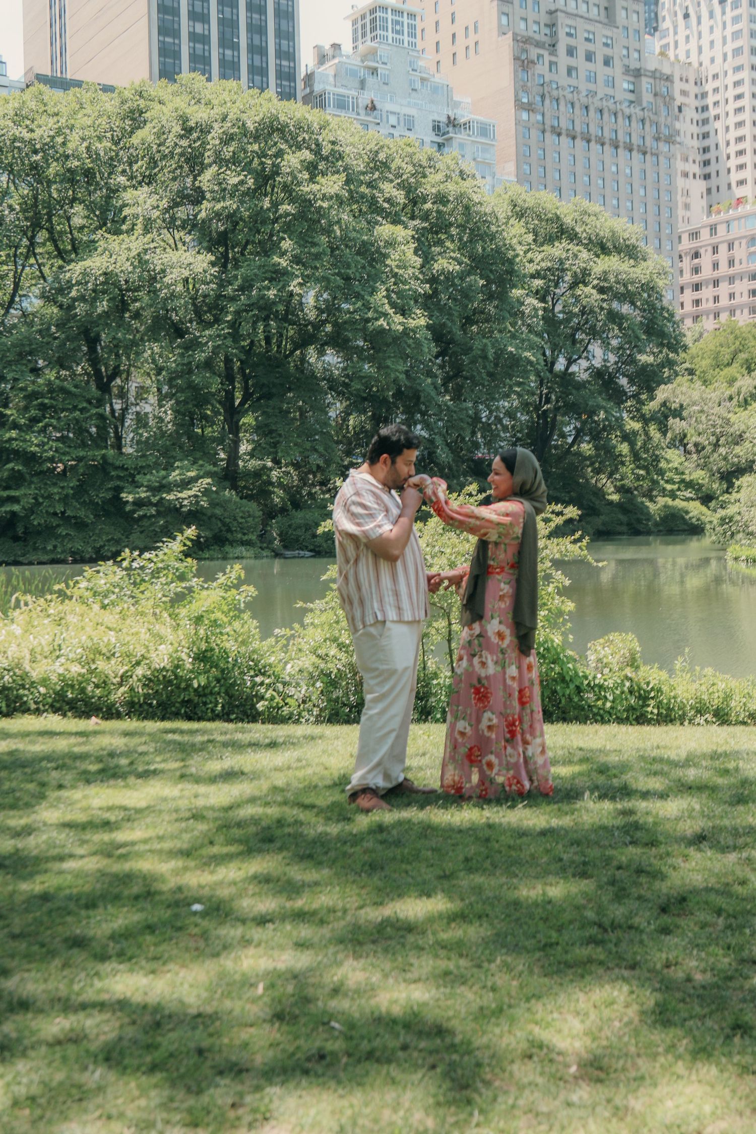 Couple in a romantic moment on a sunlit grassy lawn beside a calm pond, with lush green trees and city skyscrapers behind them.