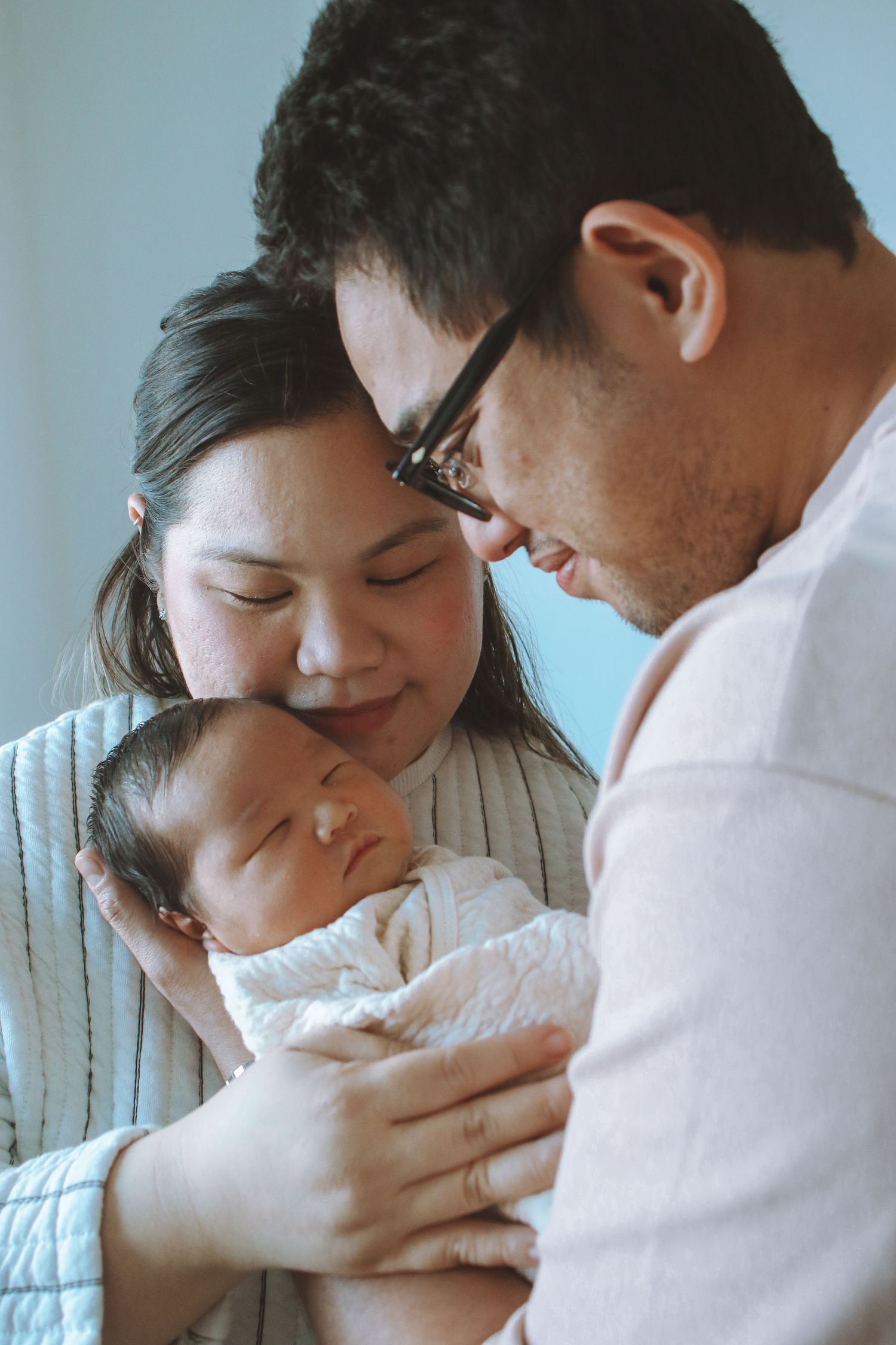 Asian parents with foreheads touching, gazing at sleeping newborn wrapped in white.