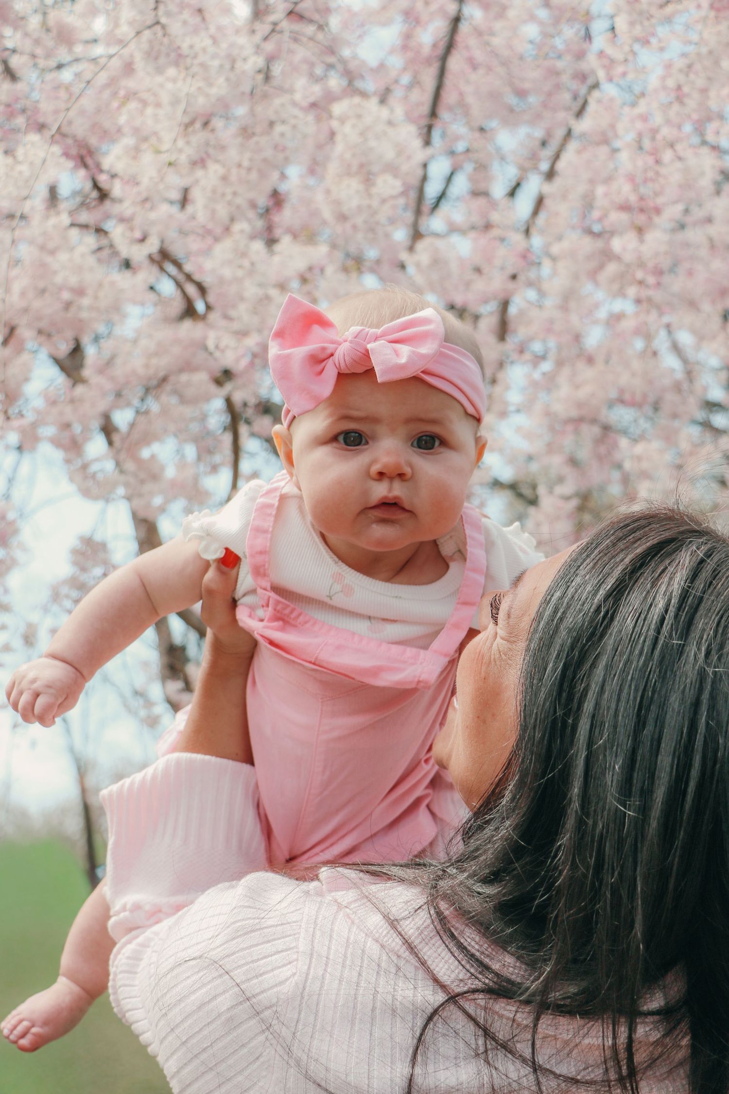 Mother lifting baby girl in pink bow headband and dress under pink cherry blossom tree.