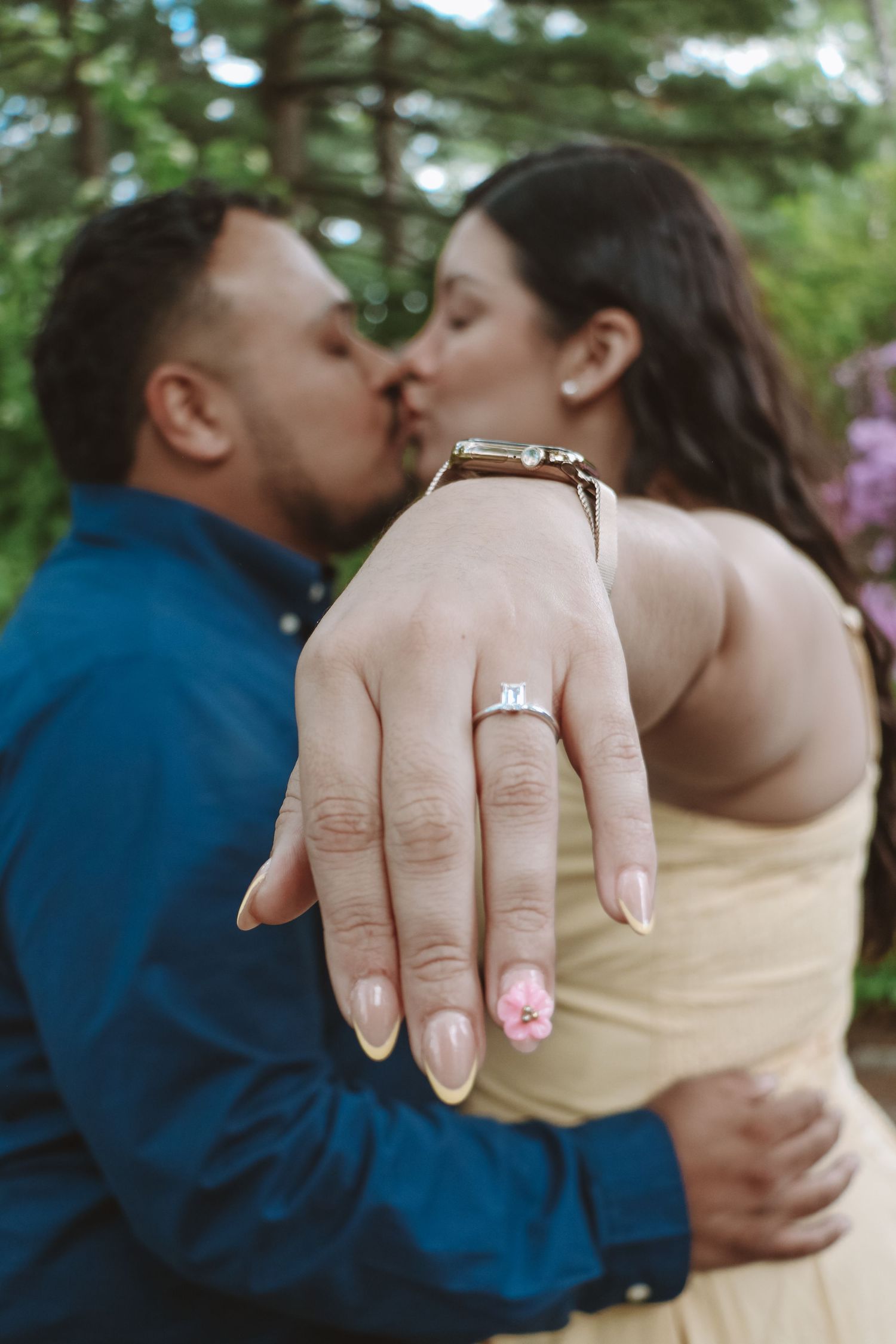 Couple kissing while showing off engagement ring with pink nail art in a garden setting.