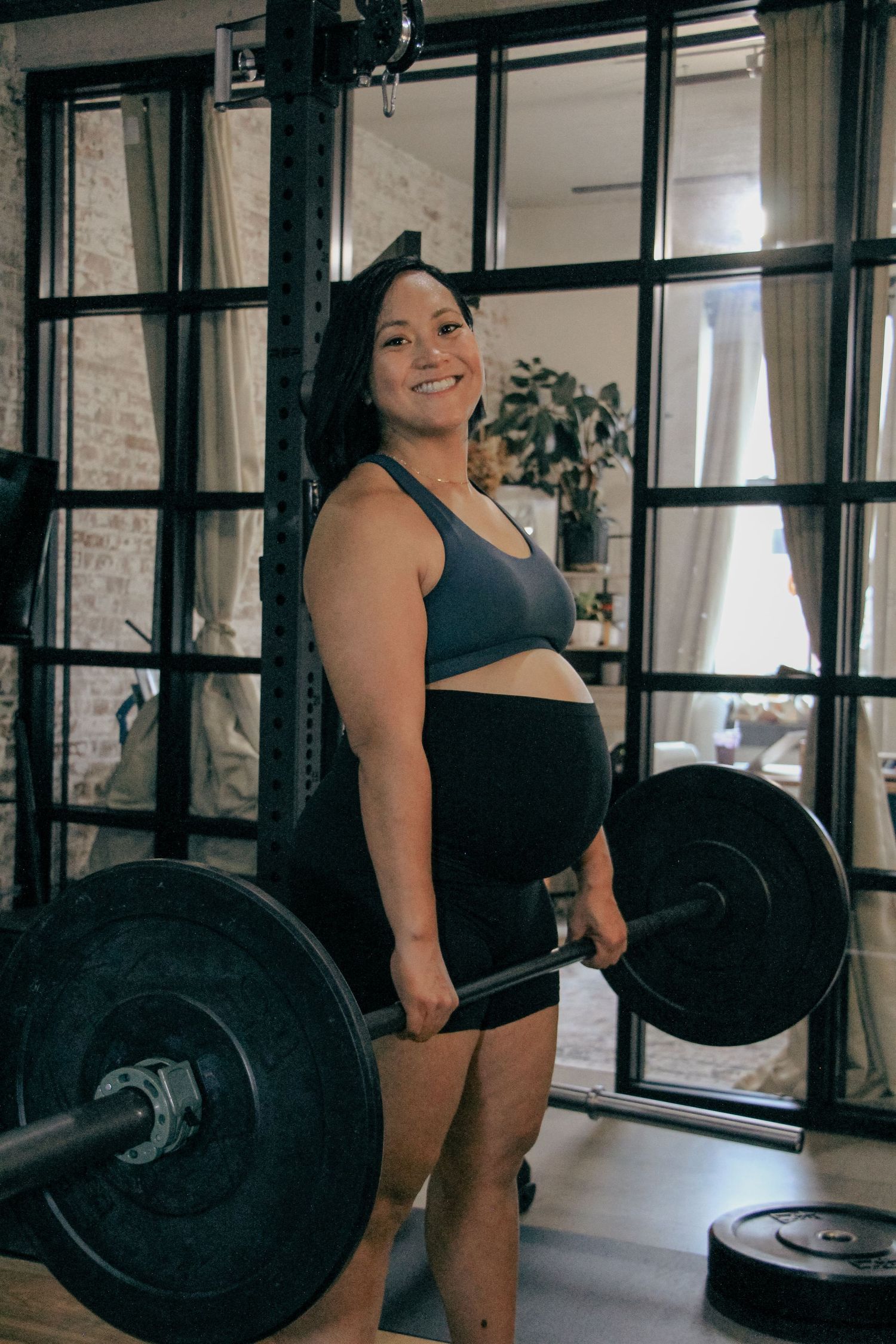 Smiling pregnant woman in a blue sports bra holding a barbell in a modern home gym.