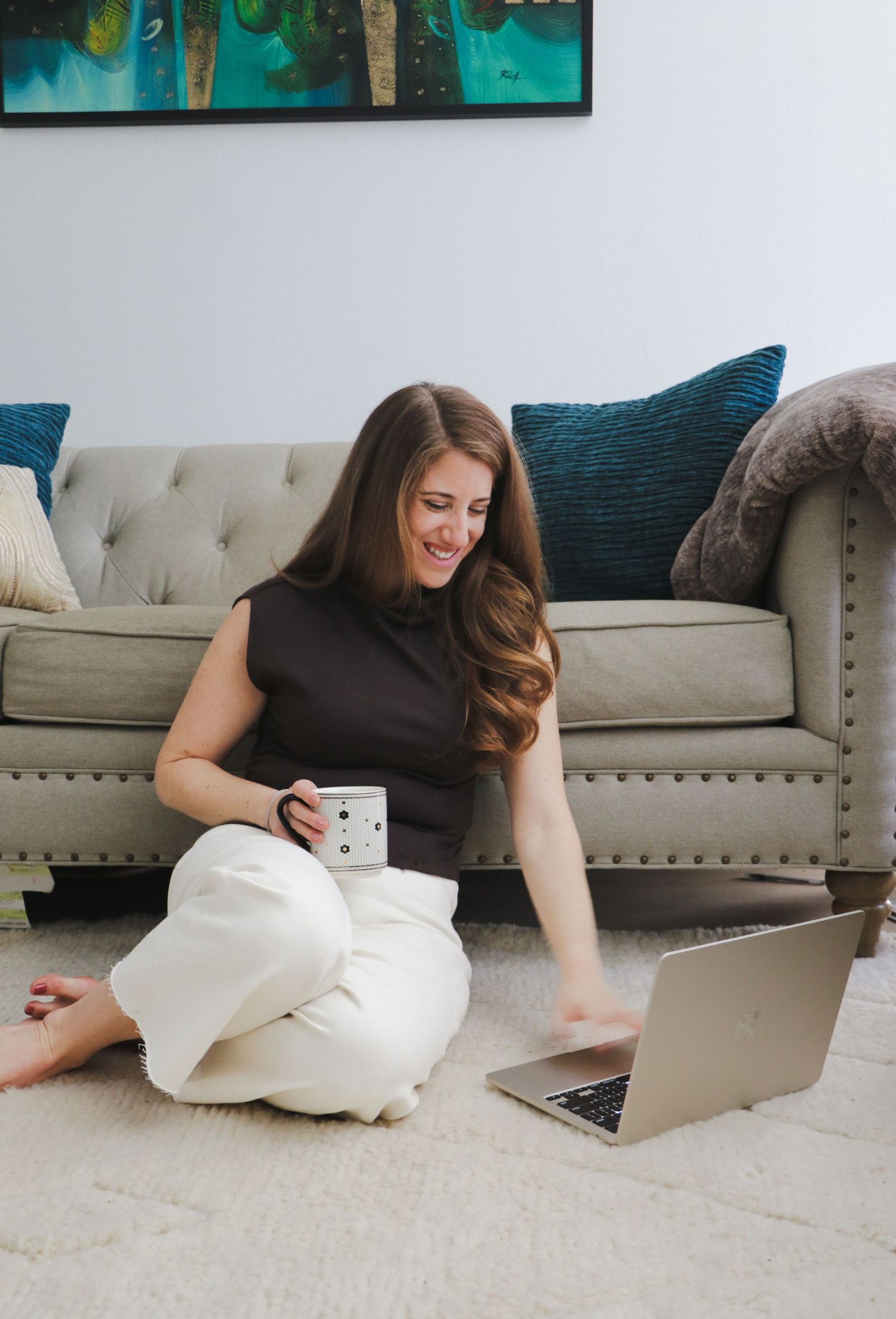 Smiling woman sitting on floor with laptop and coffee mug near gray sofa.