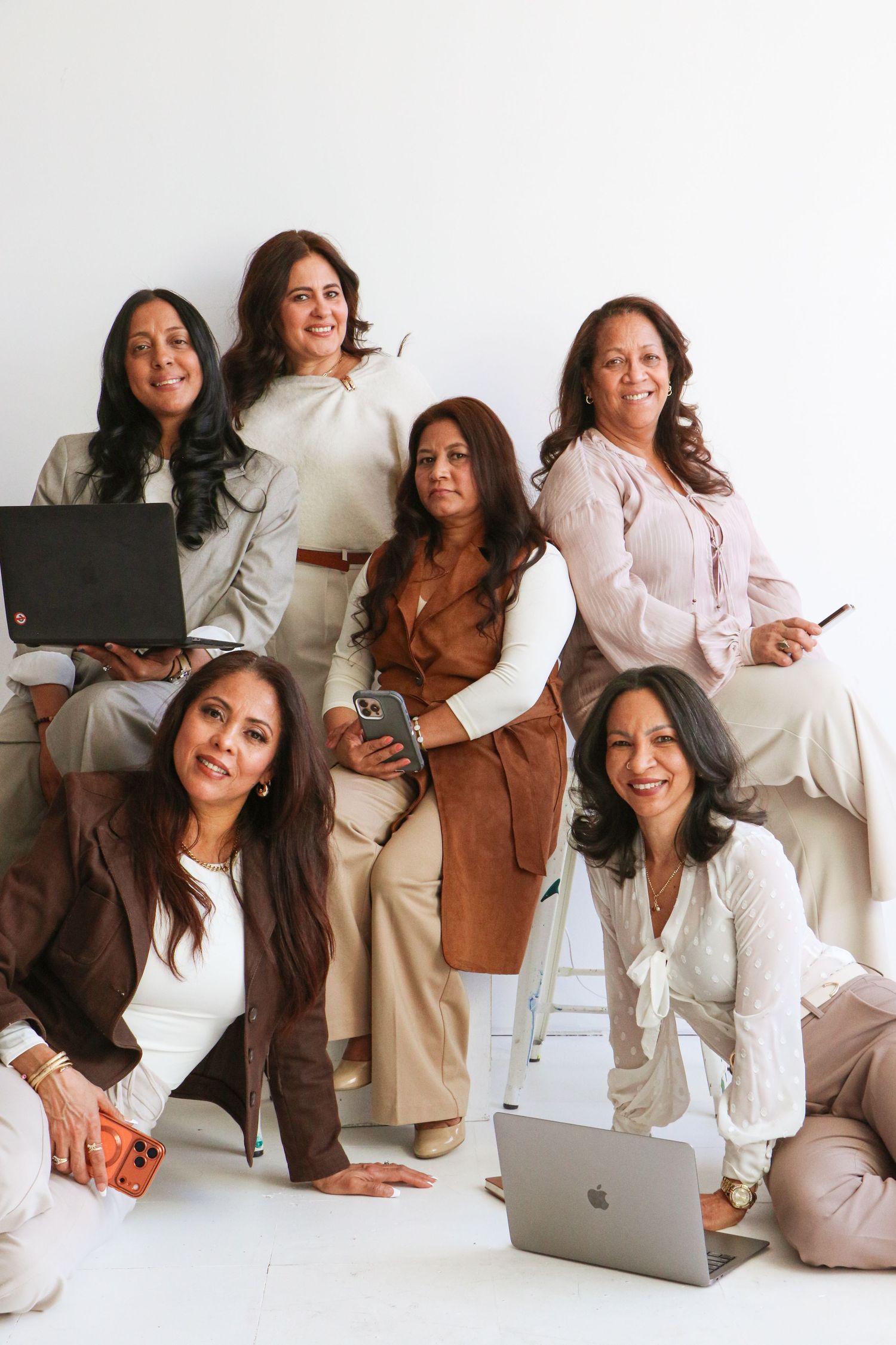 Six smiling women posing together with laptops and phones on white studio background.