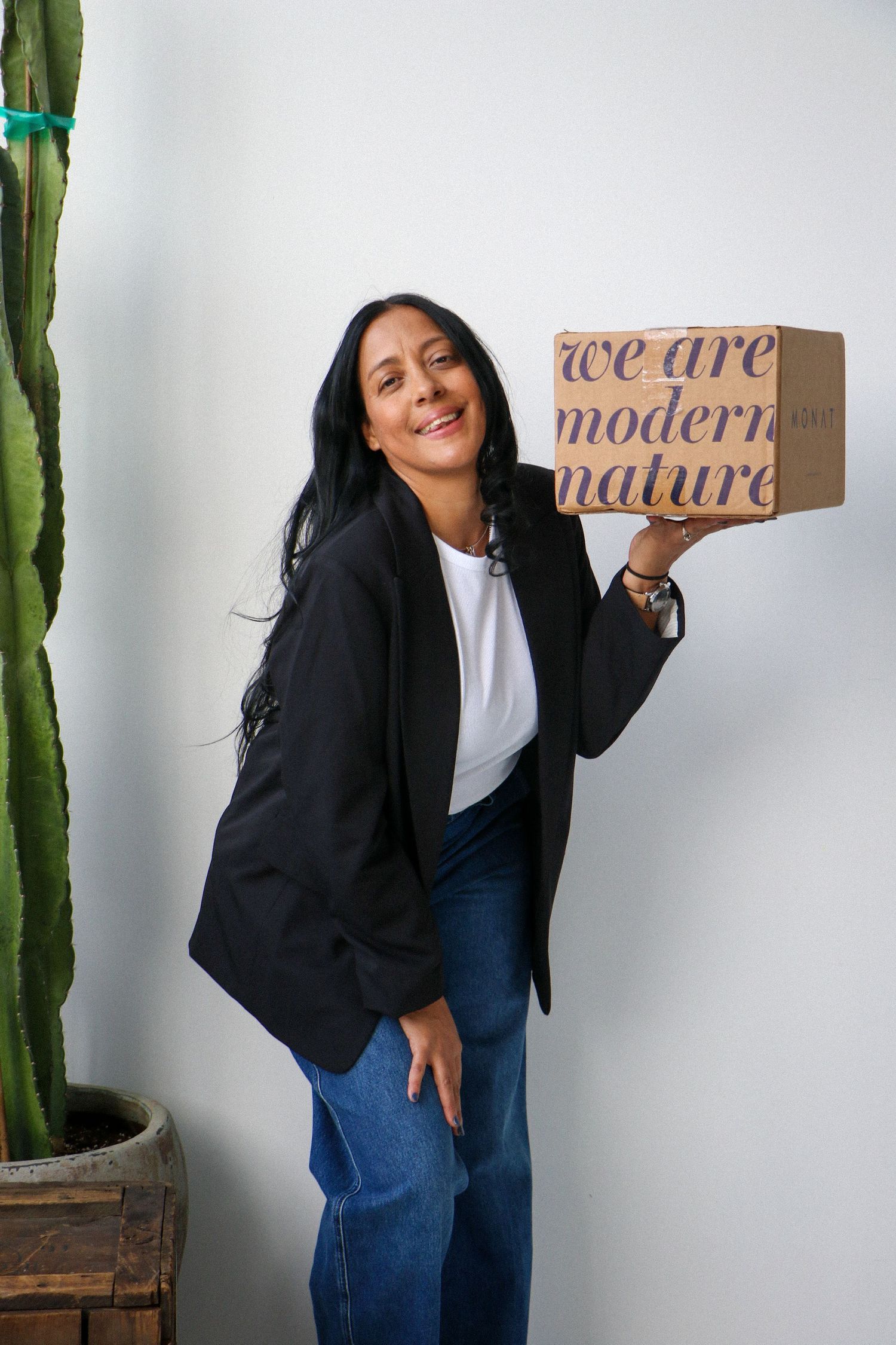 Smiling woman in black blazer holding a 'We Are Modern Nature' branded cardboard box next to a cactus.