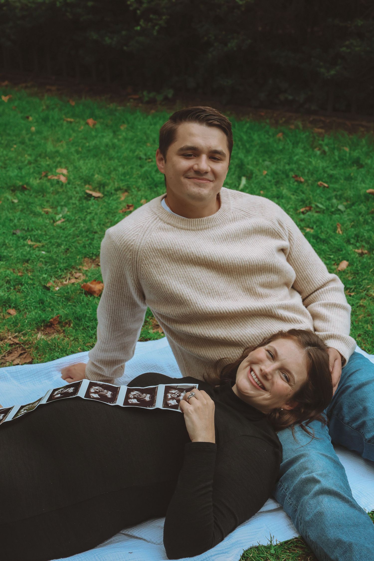 Smiling couple relaxing on a white blanket in a park as a pregnant woman holds ultrasound photos across her belly.