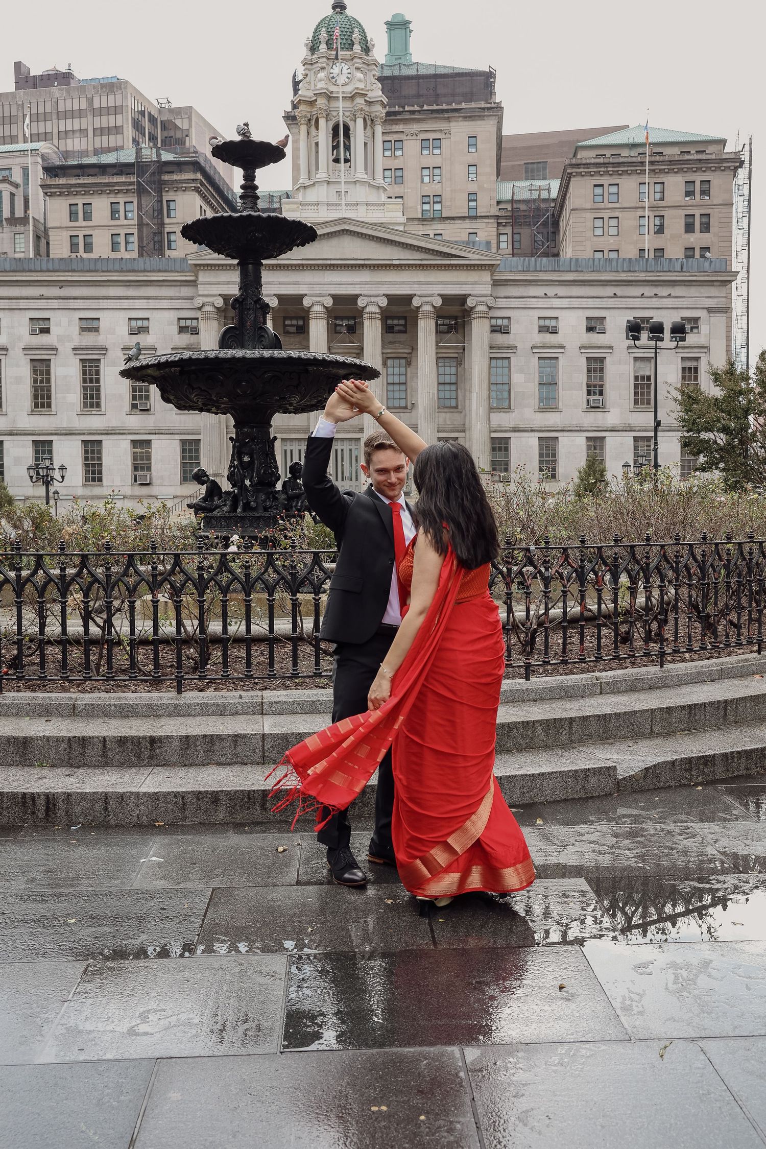 Couple dances beside a black fountain on a wet plaza, the woman twirling in a red sari before a grand neoclassical building.