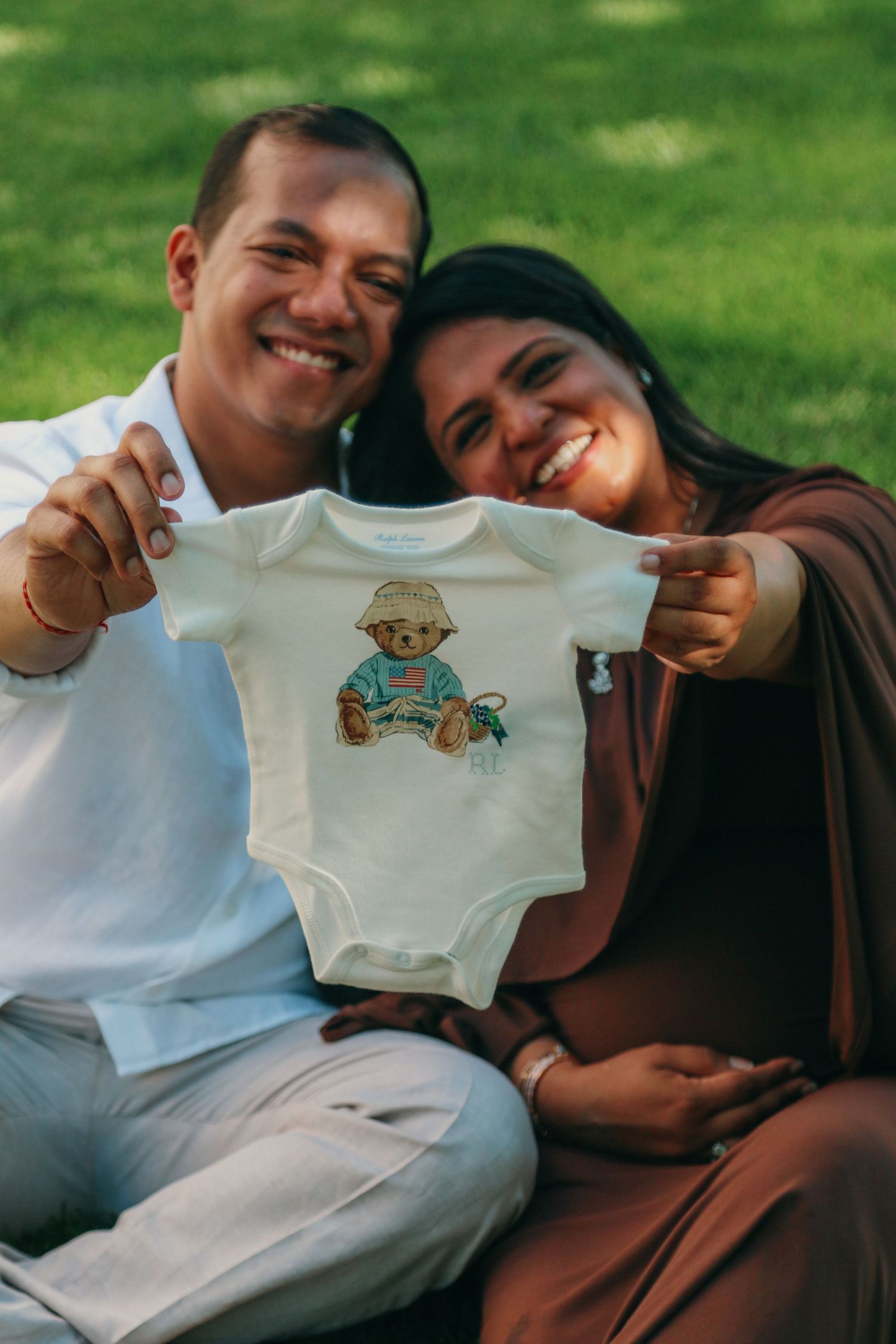 Happy couple sitting on grass holding up a white baby onesie with a bear print, announcing their pregnancy.