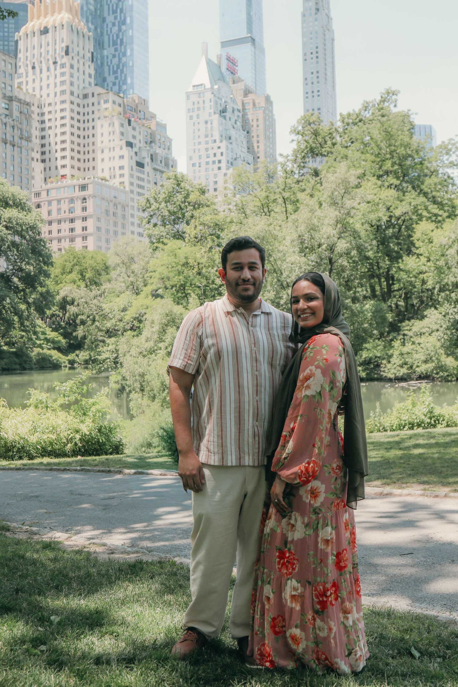 Couple posing in Central Park with lush greenery and NYC skyscrapers in the background on a sunny day.