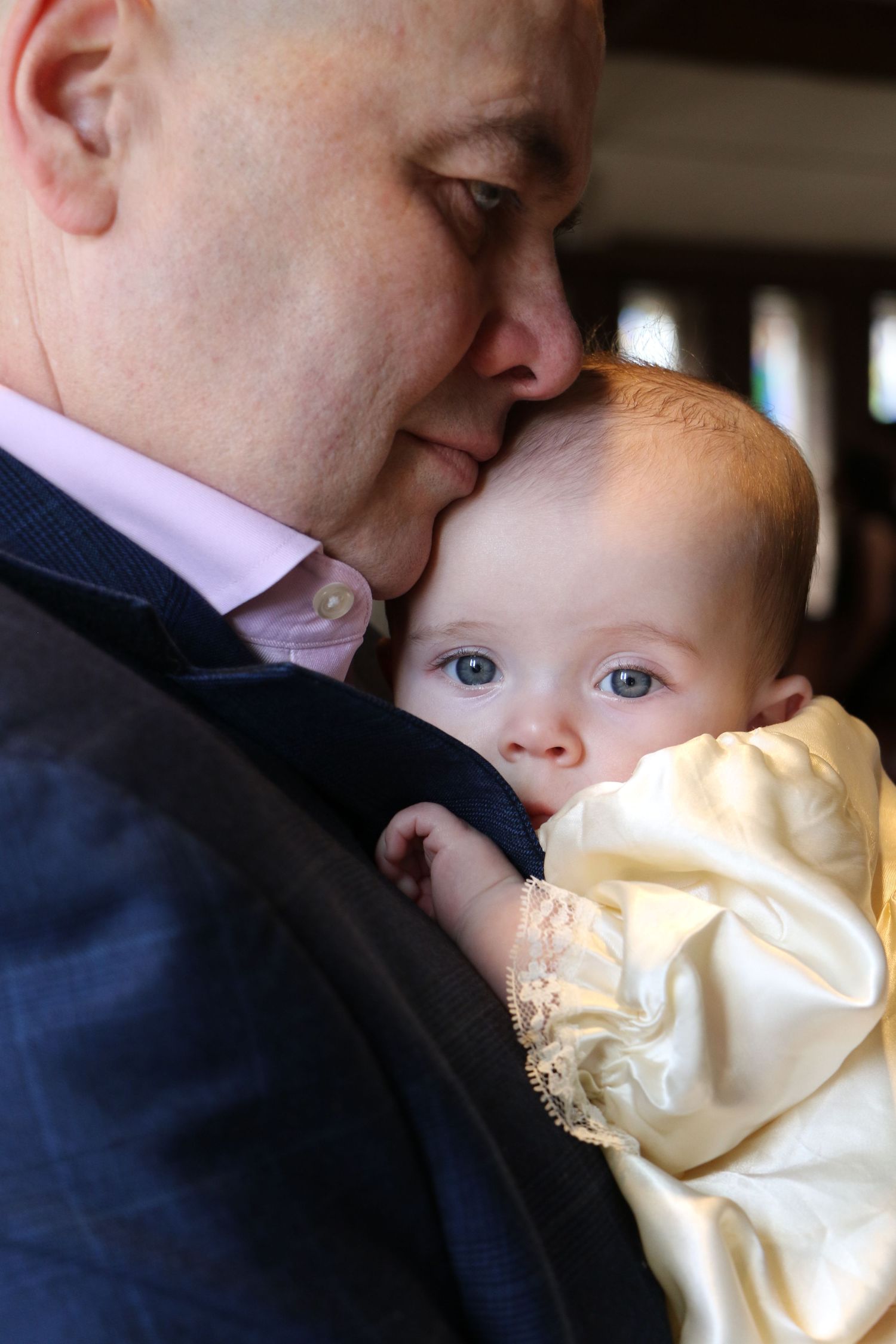 A father tenderly nuzzles his blue-eyed baby dressed in a cream christening gown at a baptism.