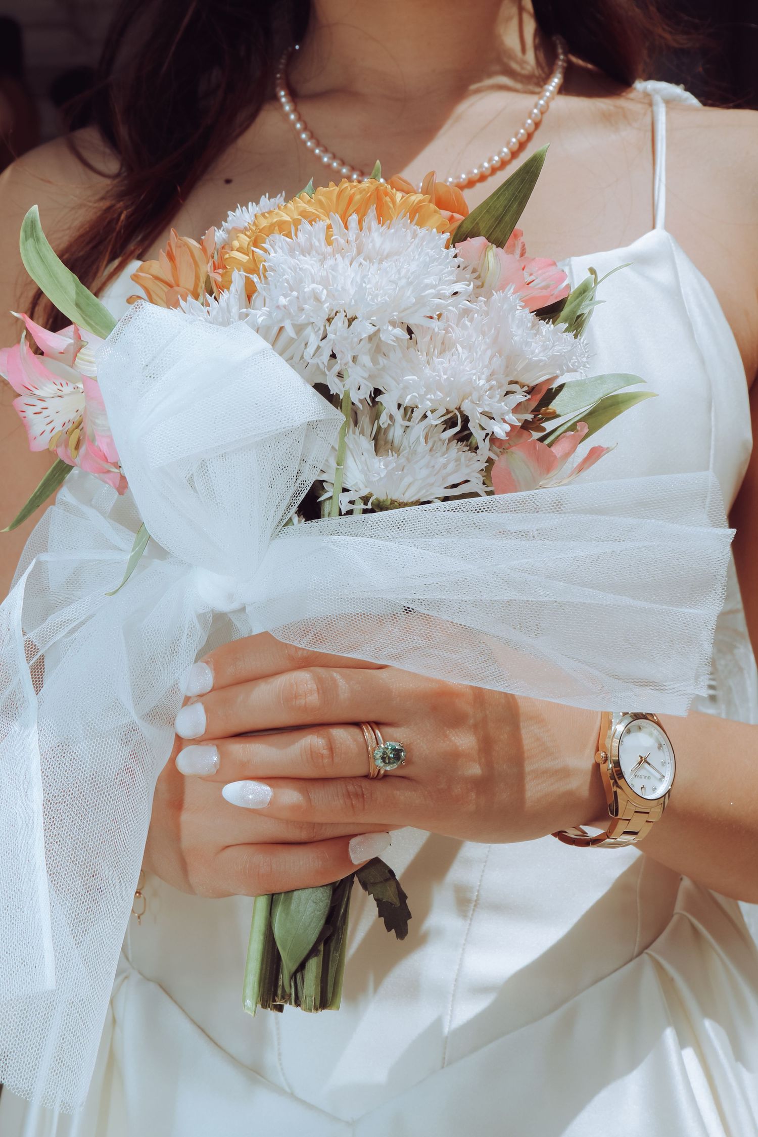 Bride in white dress holding a wedding bouquet with orange, white, and pink flowers, wearing a gold watch and green gemstone ring.