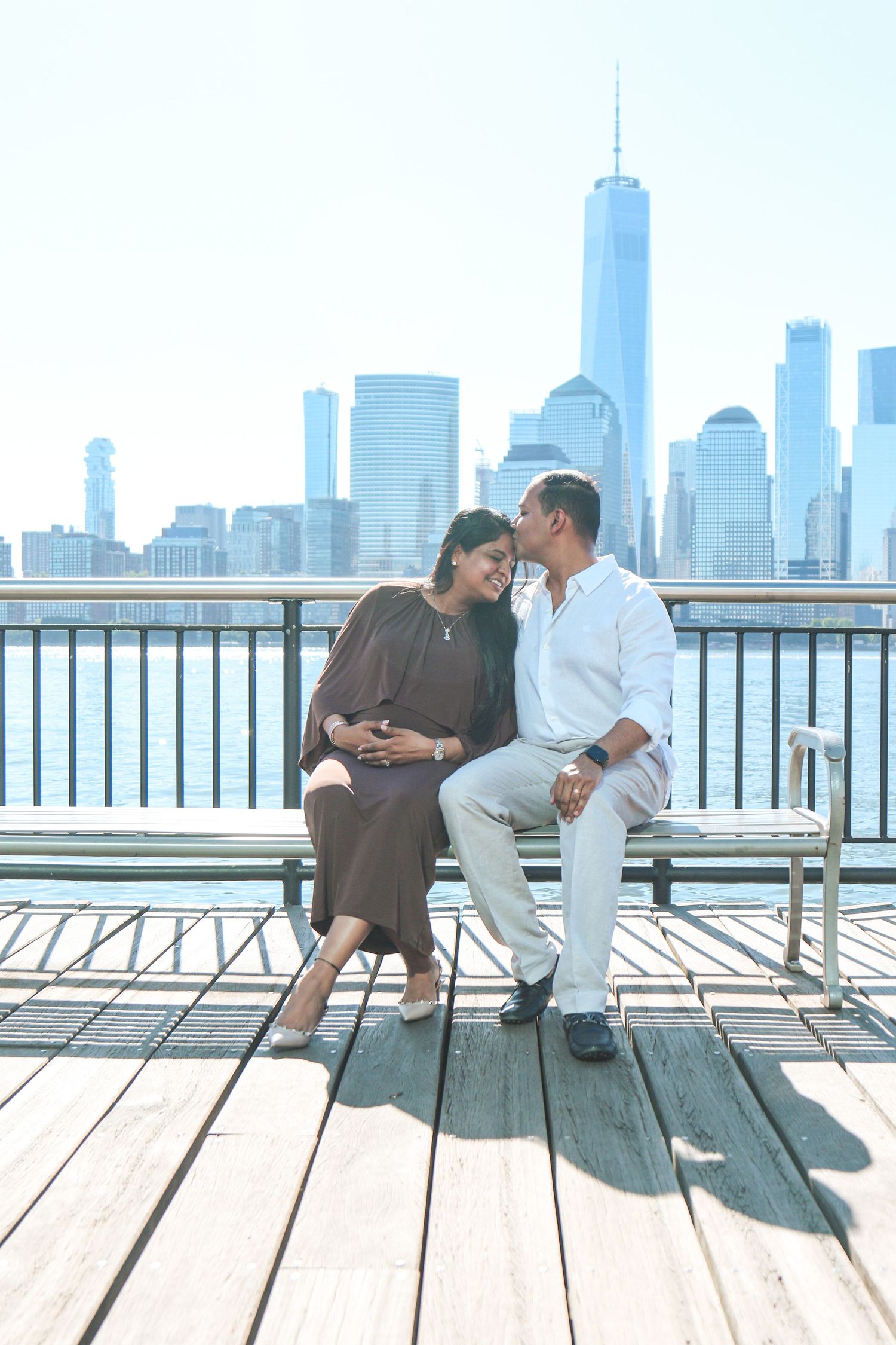 Couple sharing a romantic moment on a waterfront bench with NYC skyline and One World Trade Center in background.