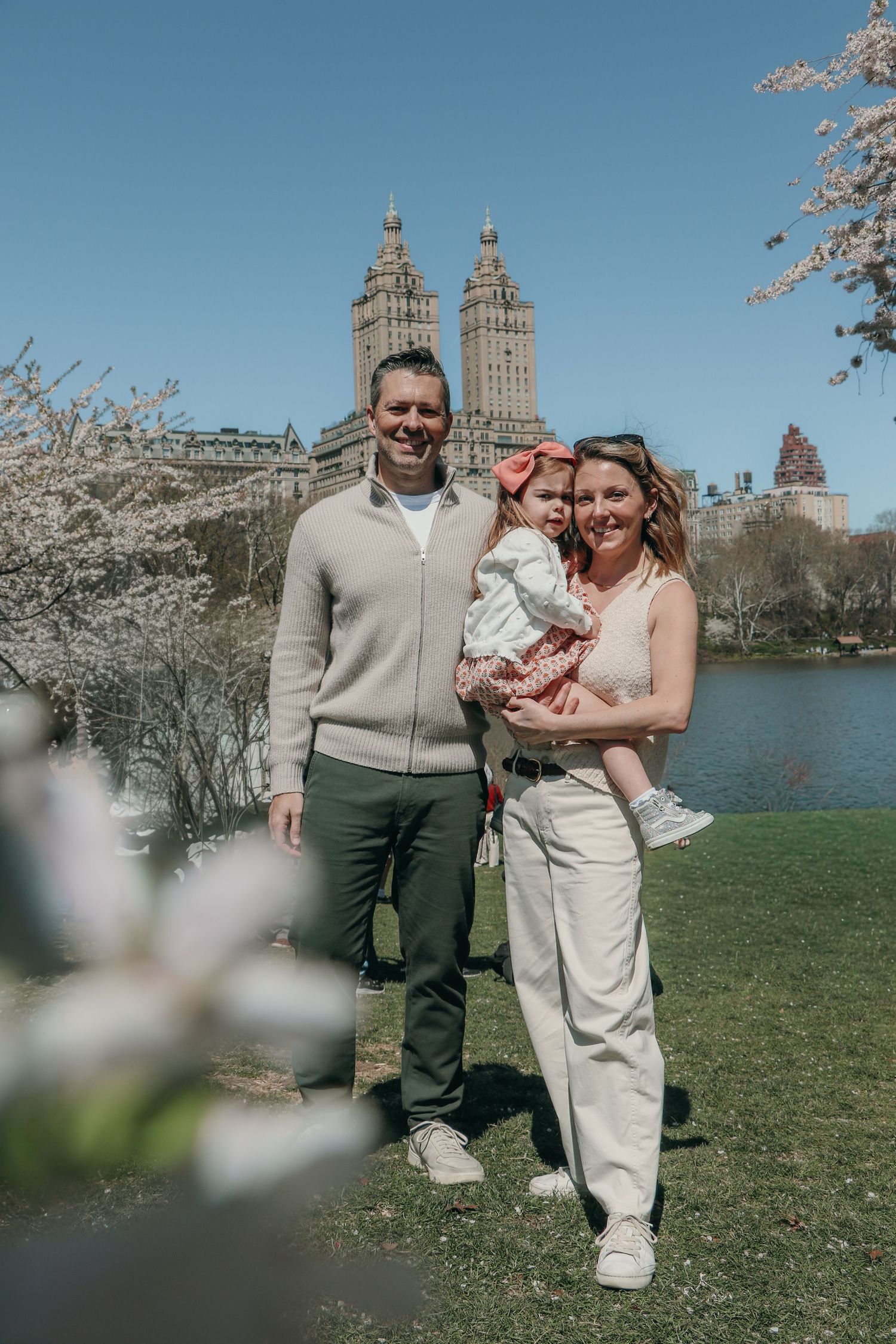 Smiling family holds young daughter among blooming cherry trees by Central Park lake.