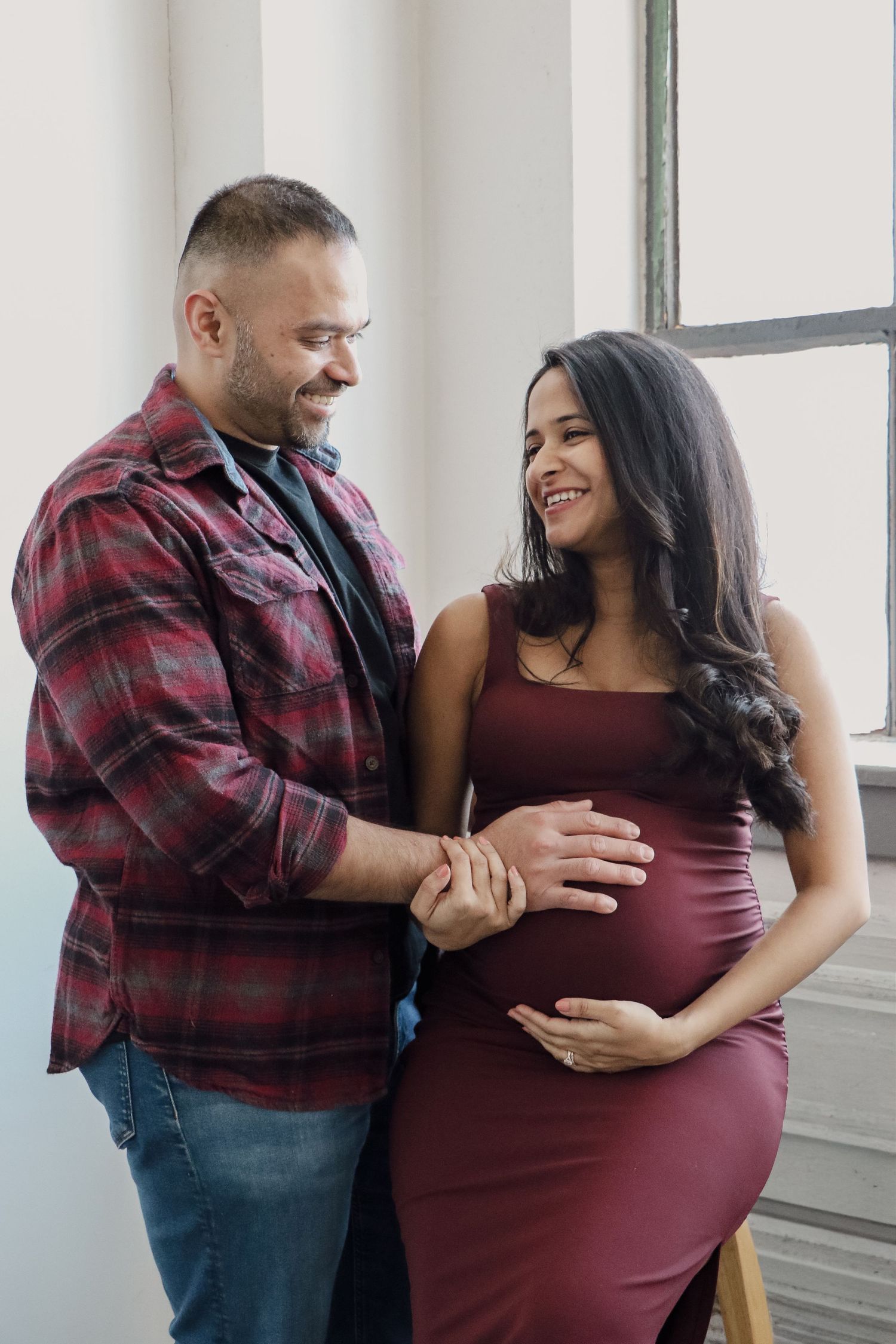 Happy couple touching pregnant belly indoors, man in plaid shirt, woman in burgundy maternity dress.