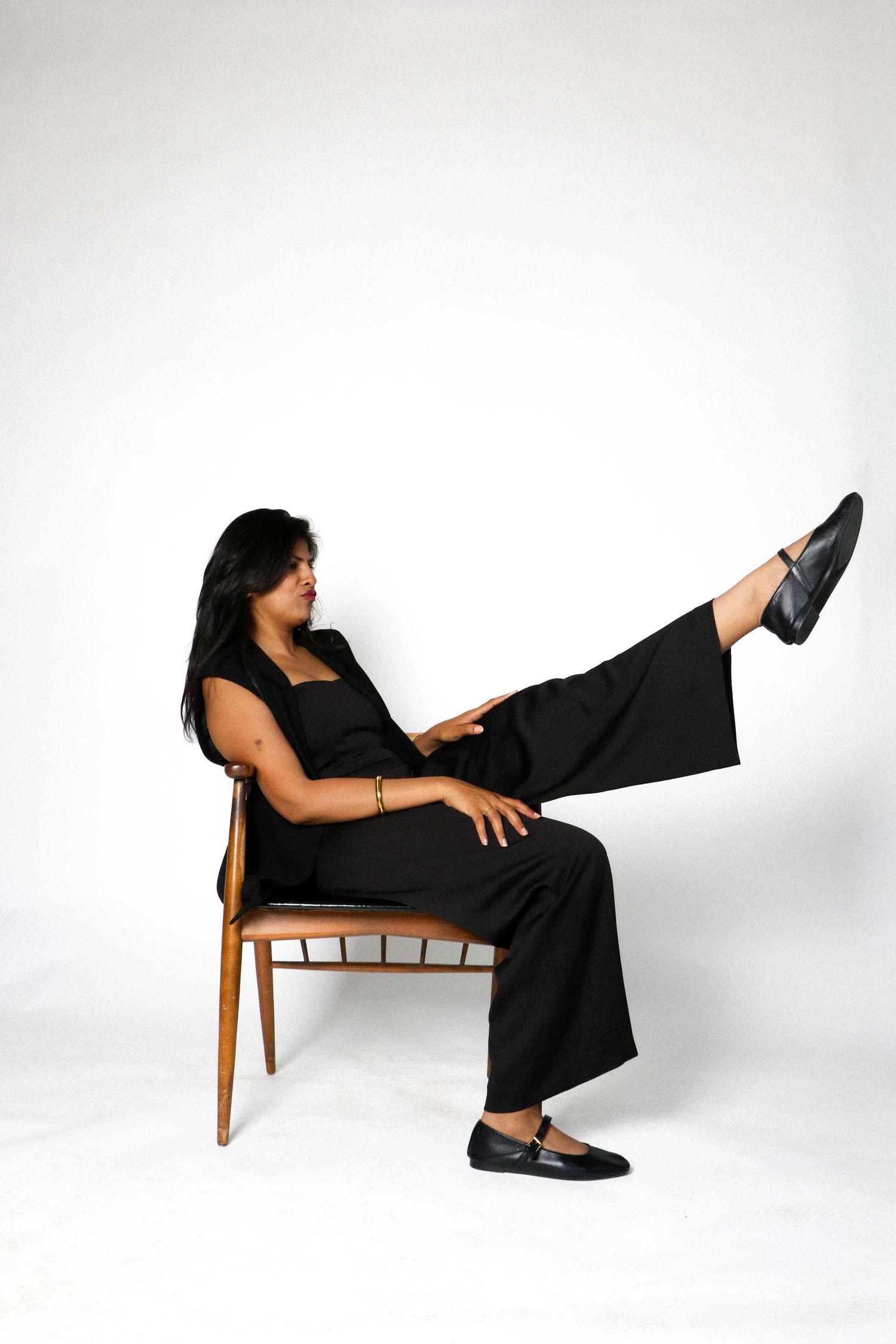 Woman in black jumpsuit sitting on wooden chair with one leg raised, posing confidently against white background.