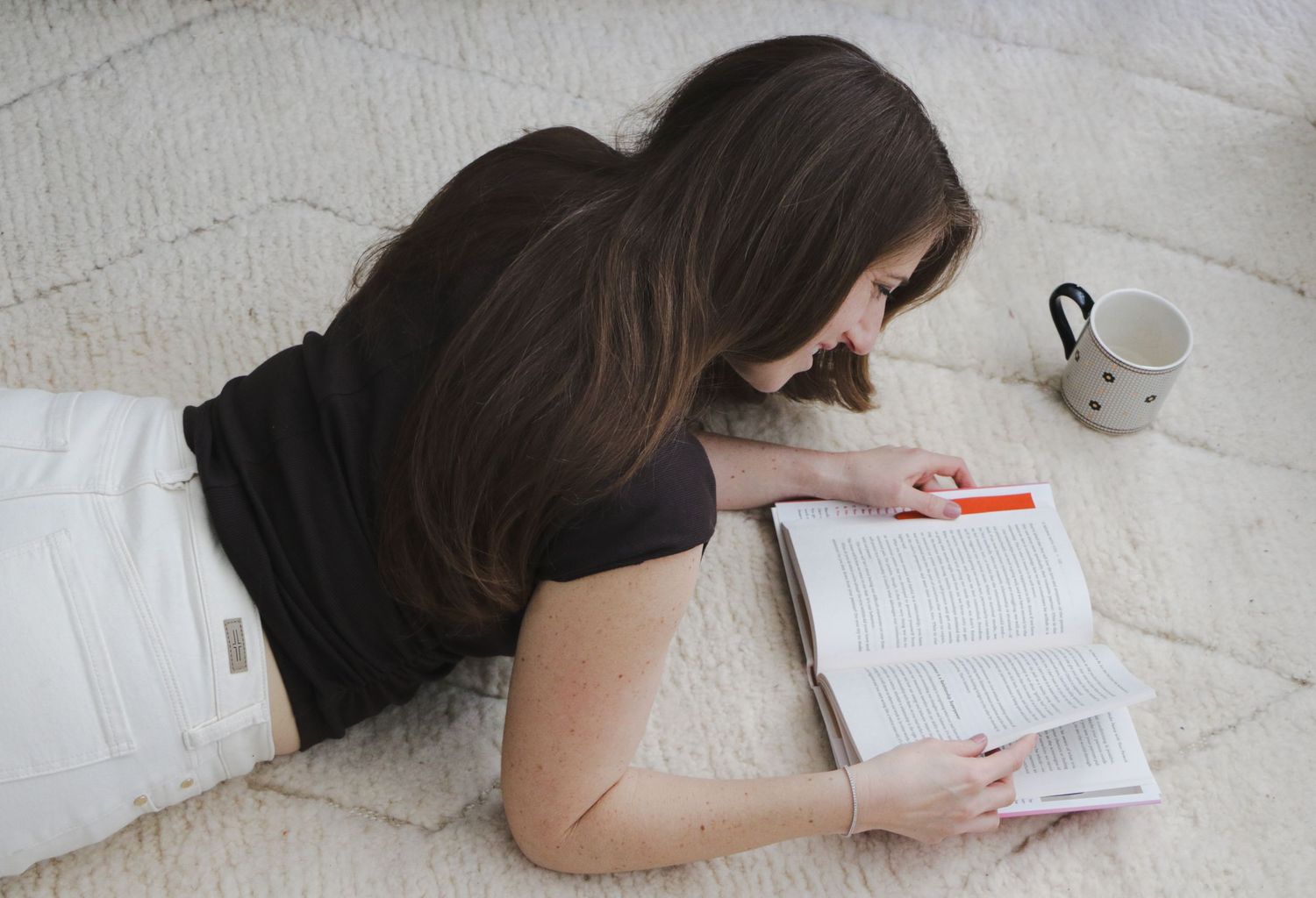 Woman lying on carpet reading book with highlighter and coffee mug nearby.