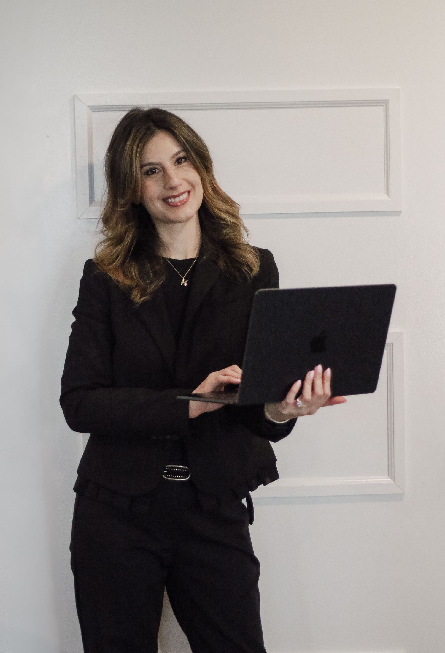 Professional woman in black blazer holding laptop against white paneled wall.